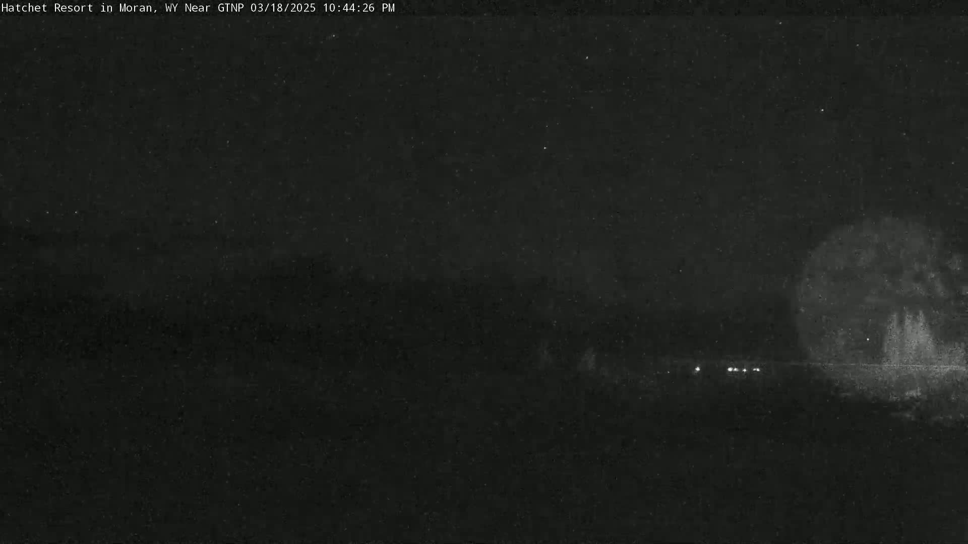 A nighttime, low-light view shows a dark landscape with a few faint lights in the distance and a brighter area that may be trees to the right.