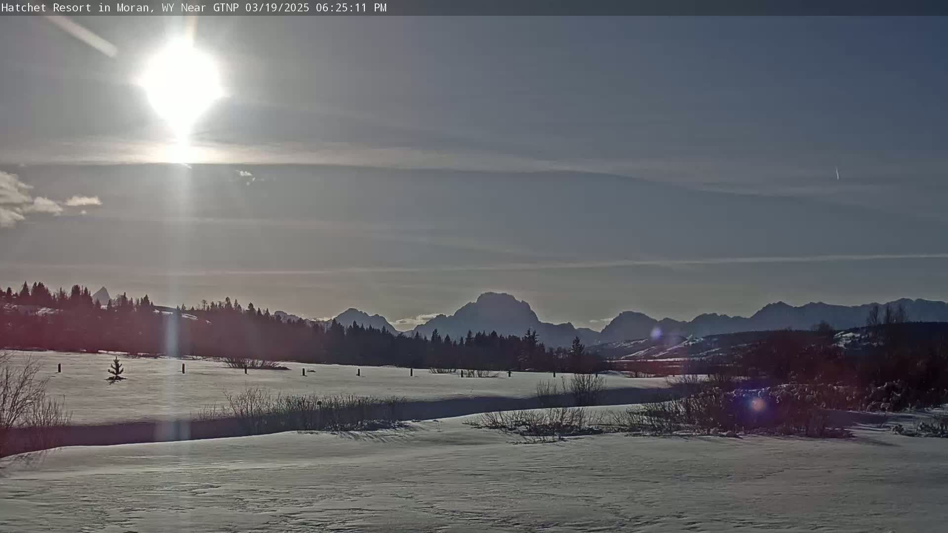 A bright sun shines in a mostly clear sky over a snow-covered landscape with a line of mountains in the distance.