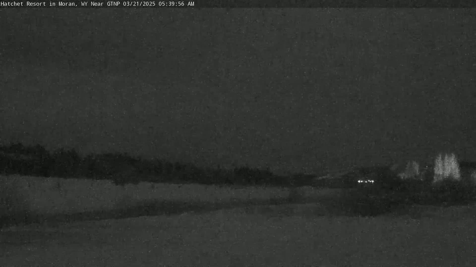 A dark, nighttime scene shows a flat, snow-covered landscape with a line of trees in the distance and a few faint lights visible near the horizon.