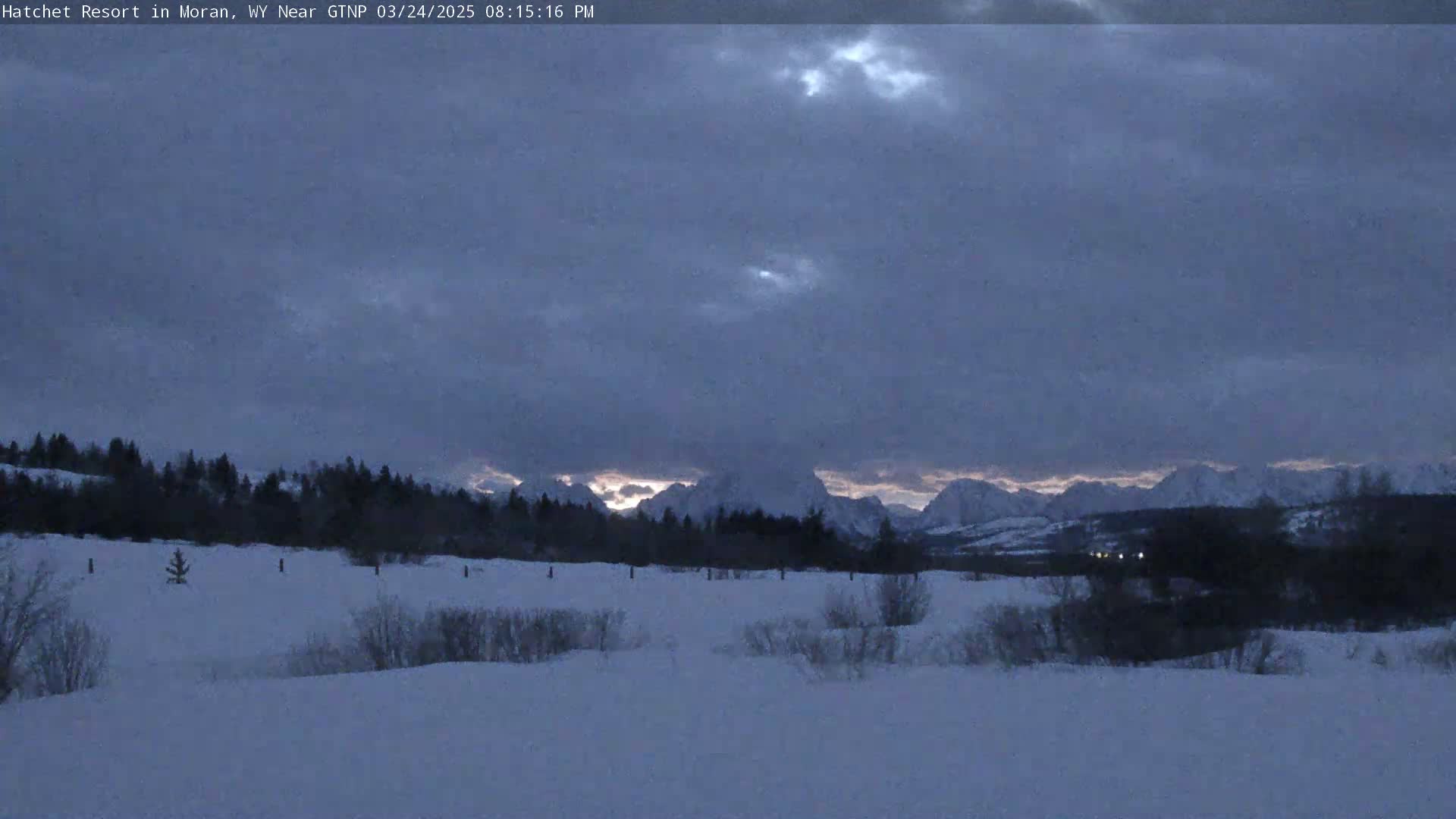 A snow-covered field stretches toward a line of dark trees and snow-capped mountains under a dusky, cloudy sky.