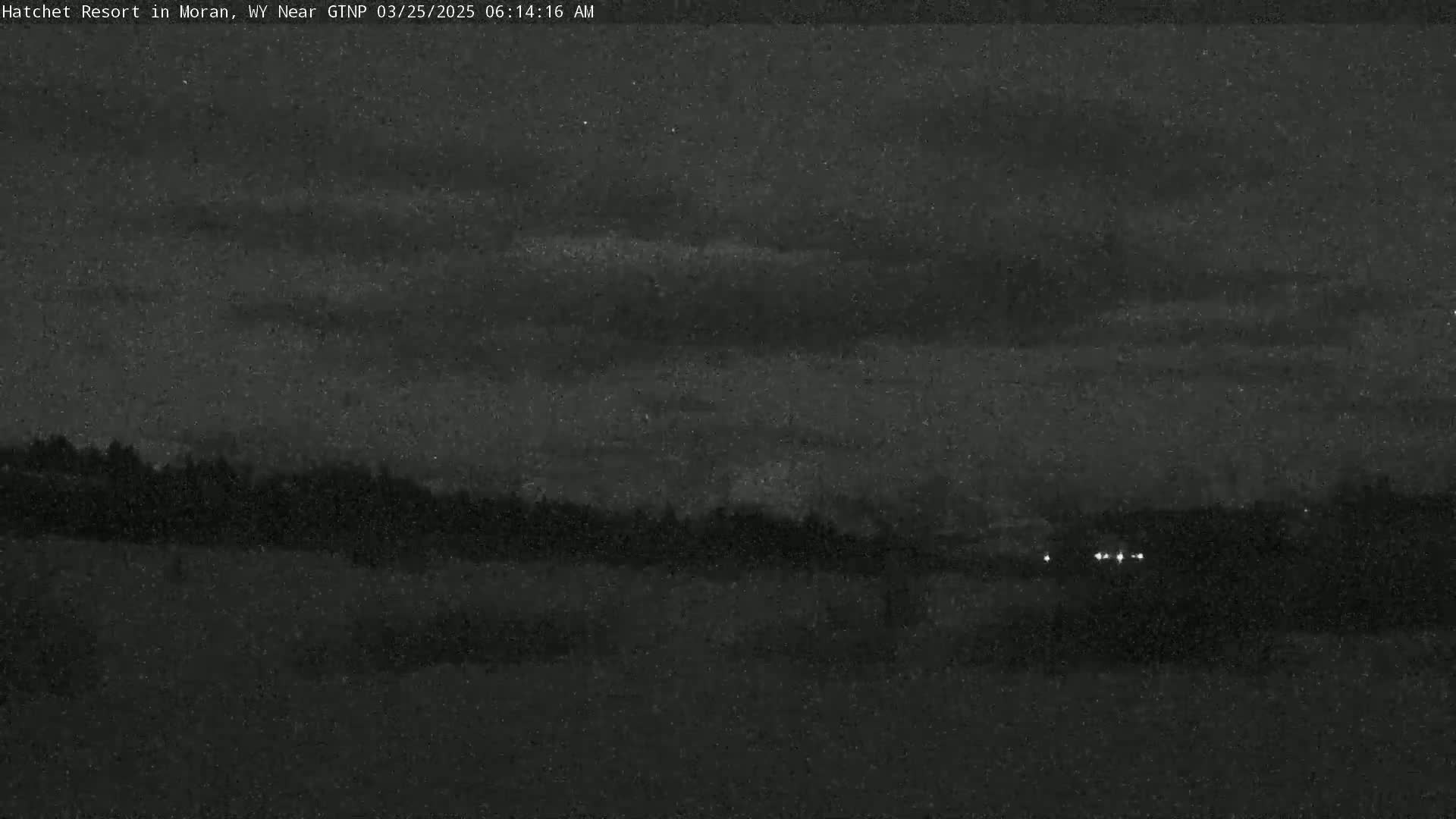 A nighttime, low-light view shows a dark, flat landscape with a line of trees or shrubs in the middle distance, and a few distant lights visible near the horizon under a mostly cloudy sky.