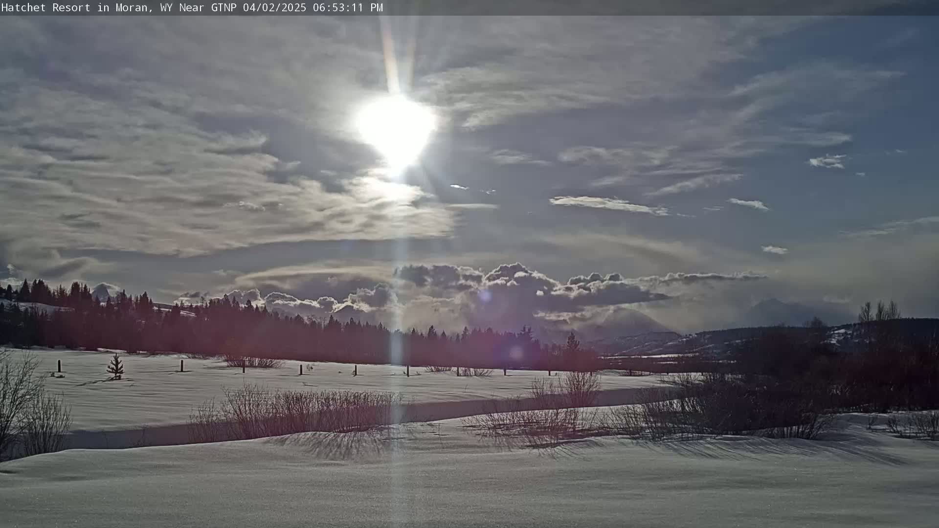 A bright sun shines through a partly cloudy sky over a snow-covered field and a line of dark trees, with mountains visible in the distance.