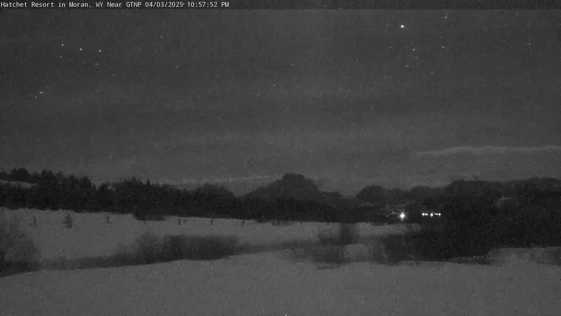 A nighttime, snowy landscape shows a dark, snow-covered field, trees, and distant hills under a clear, starry sky.