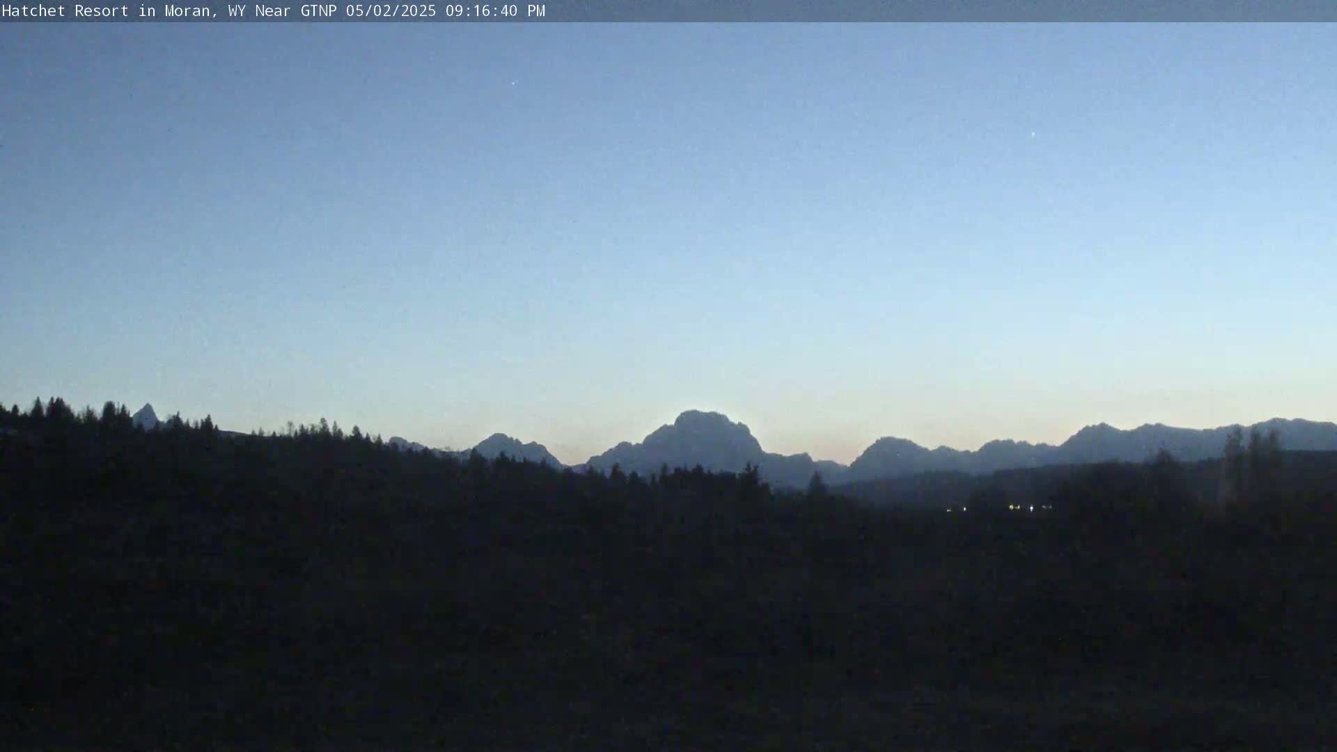 A dark, silhouetted landscape of trees and mountains is visible under a clear, twilight sky.