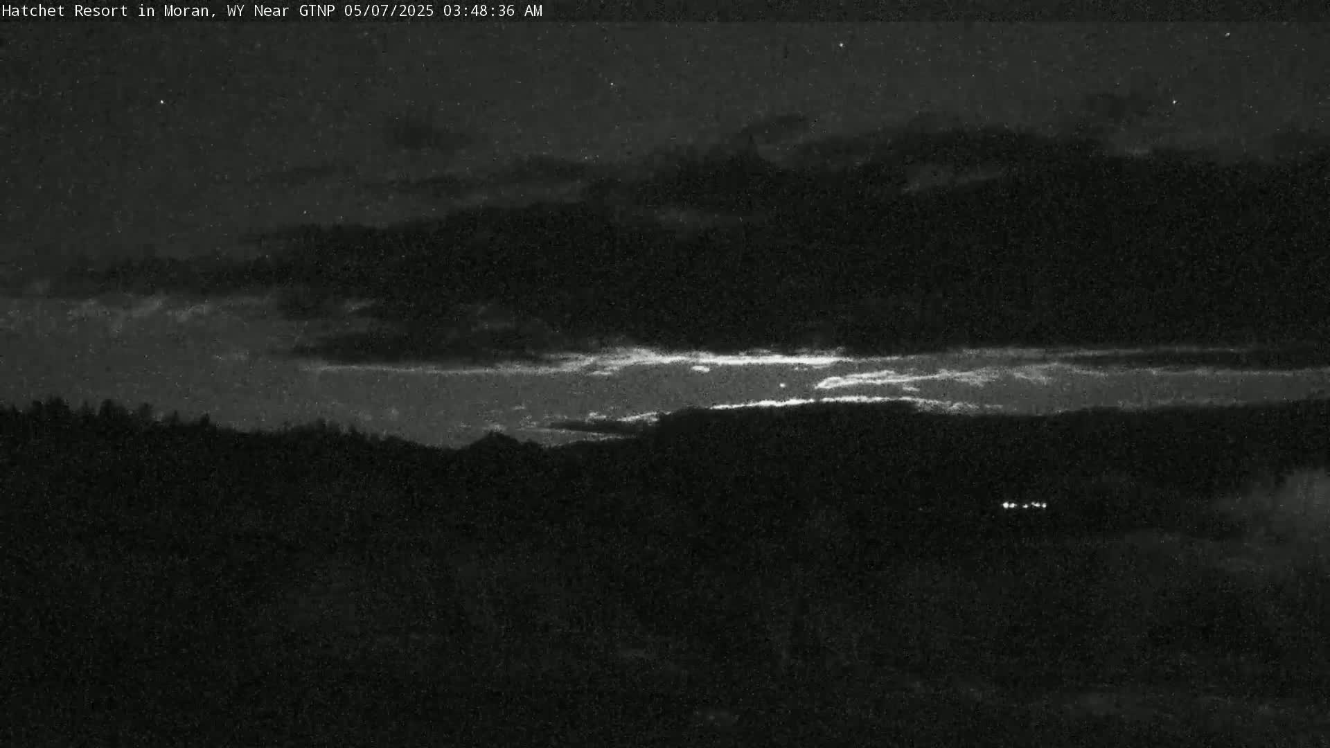 A nighttime view of a dimly lit landscape with a thin band of brighter light on the horizon under a mostly cloudy sky.