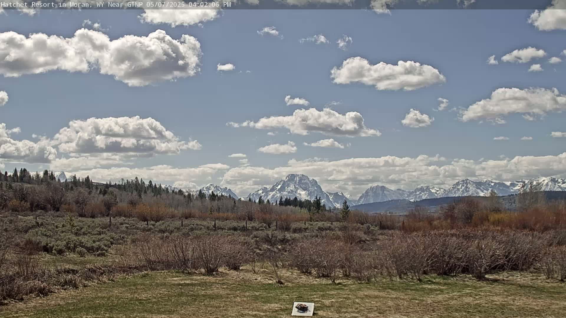 A partly cloudy day reveals snow-capped mountains in the distance, beyond a field of low-lying, brown brush.