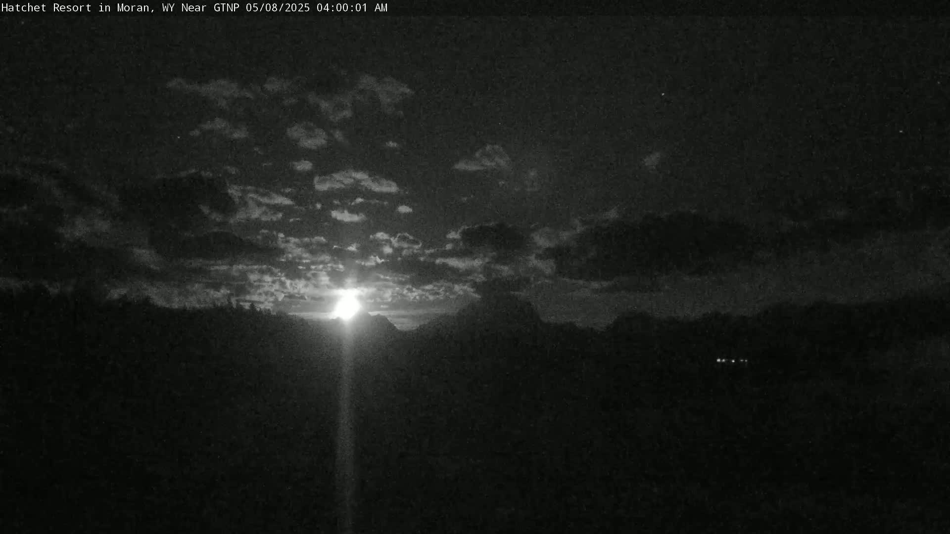 A nighttime view of a sunrise or moonrise over a dark, silhouetted landscape under a cloudy sky.