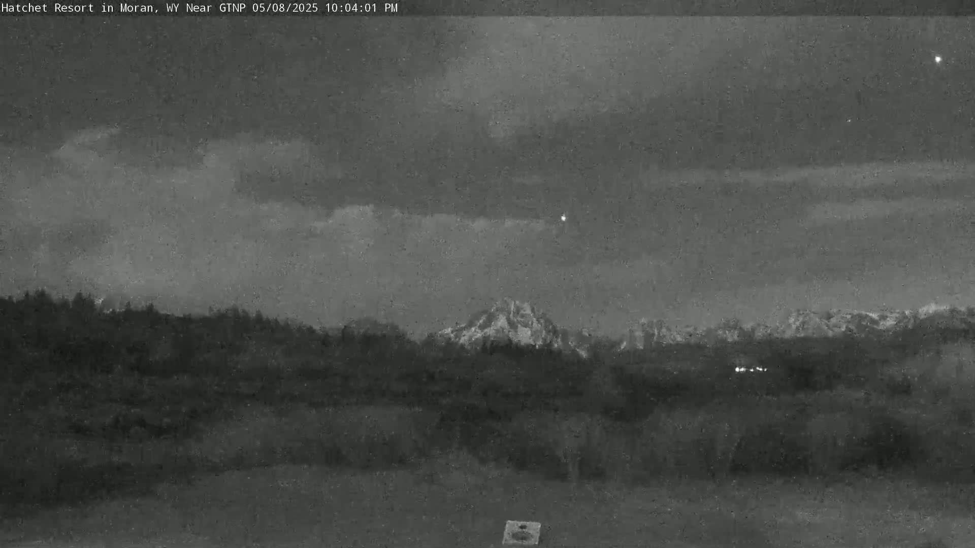 A nighttime view shows a mountain range behind a dark forest under a cloudy sky.