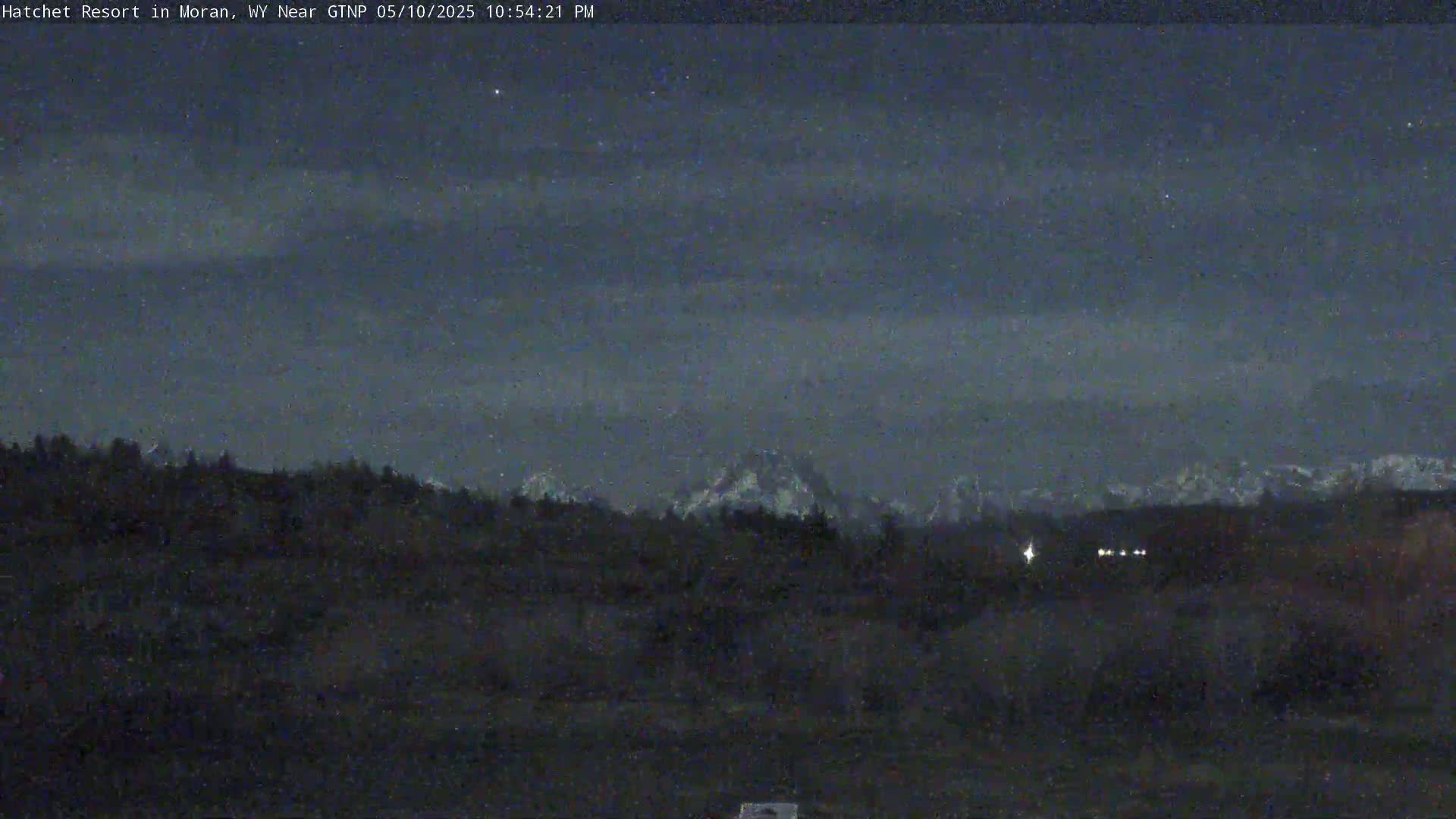 A nighttime landscape shows dark trees and mountains under a partly cloudy sky speckled with stars.