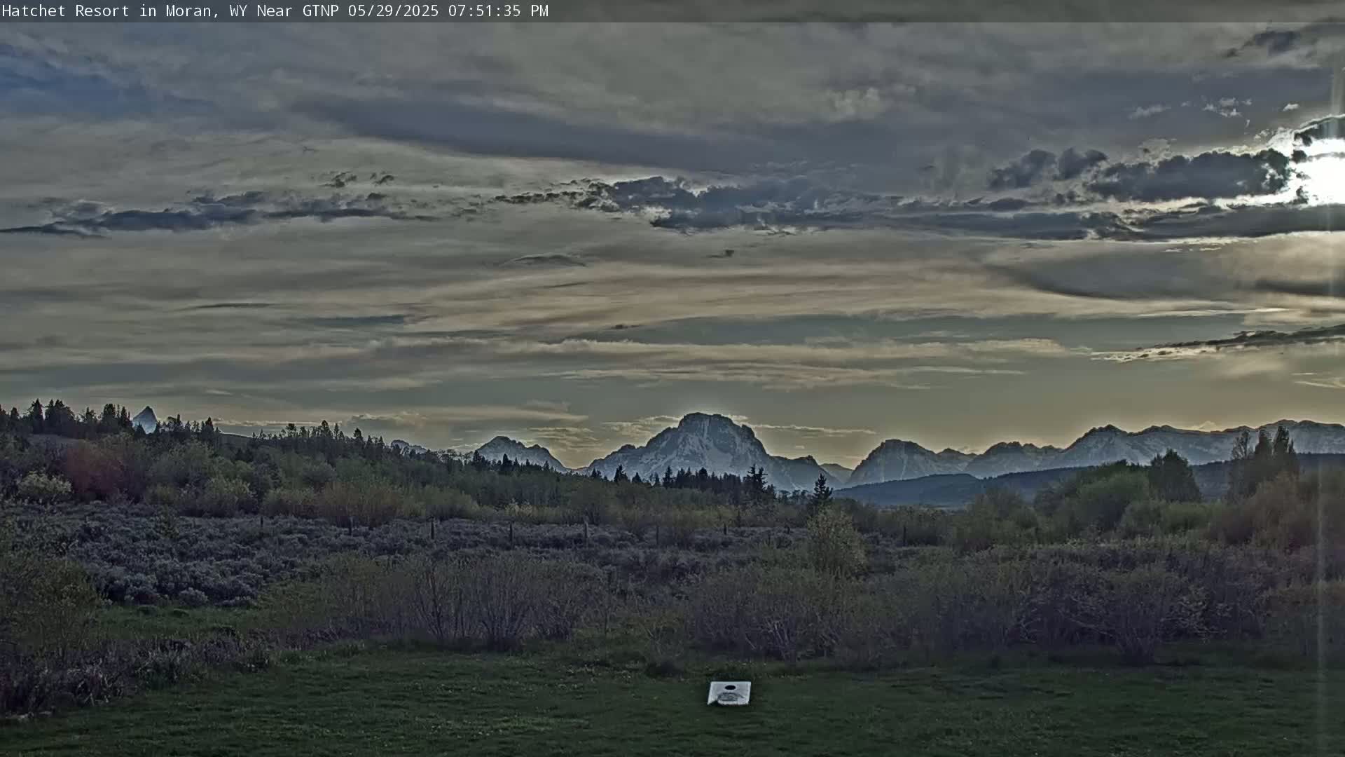 A field with low-lying vegetation and a single cornhole board sits before a mountain range partially obscured by a cloudy sky at dusk.