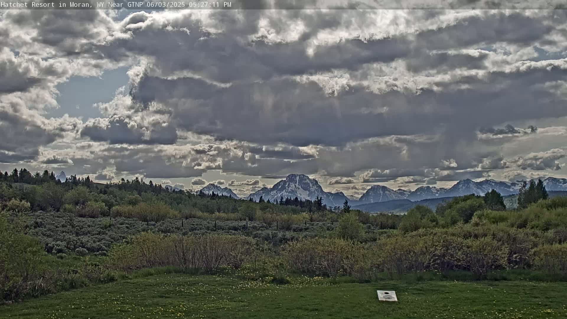 A grassy field with a cornhole board sits before a verdant landscape, which in turn precedes a partially snow-capped mountain range under a mostly cloudy sky.