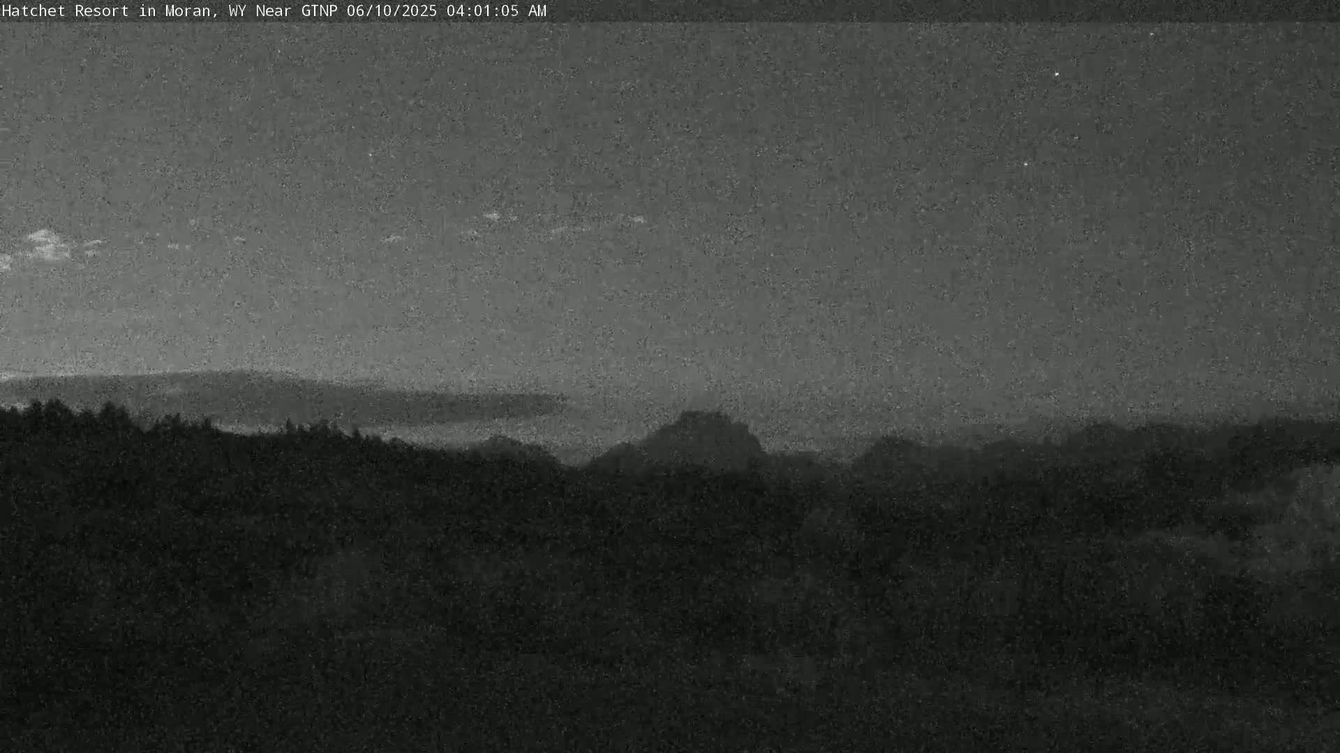 A low-light, nighttime view shows a dark landscape of distant mountains and a foreground of trees under a mostly clear sky.