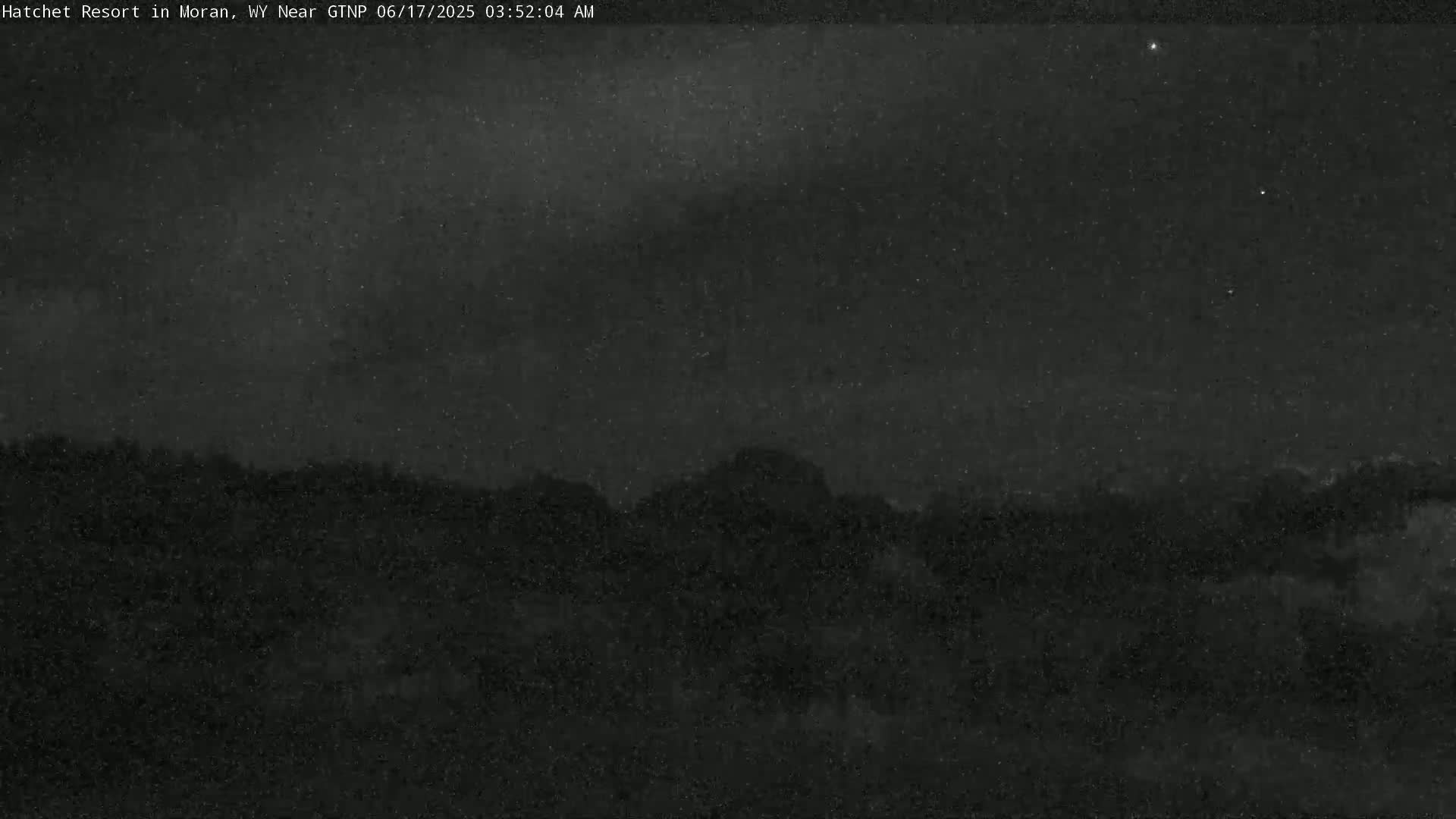 A nighttime, low-light outdoor image shows a dark, silhouetted mountain range under a mostly clear, starry sky.