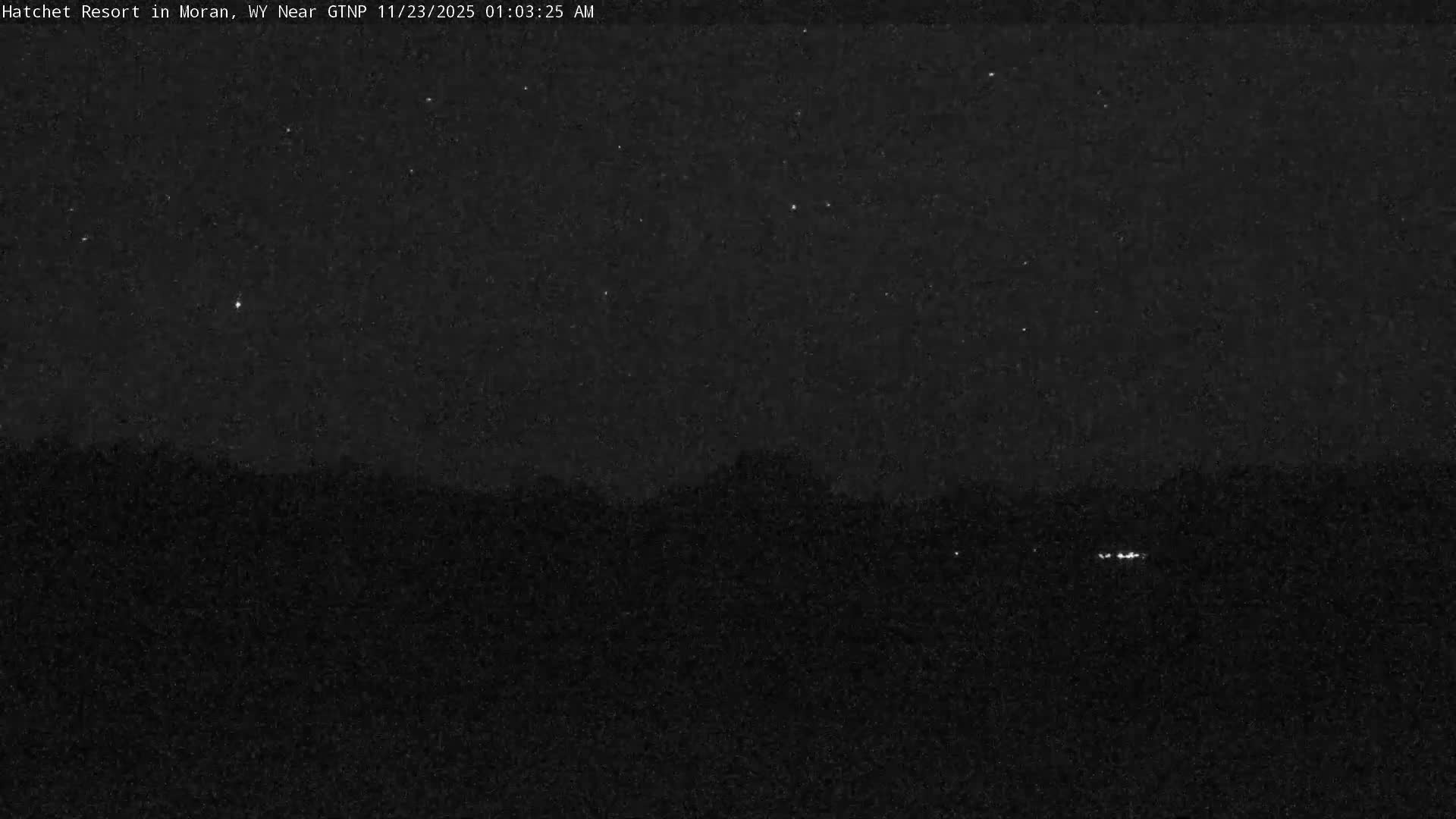 A dark, grainy nighttime image reveals a clear sky with scattered stars above a dimly visible, indistinct landscape featuring a few distant lights on the right horizon.