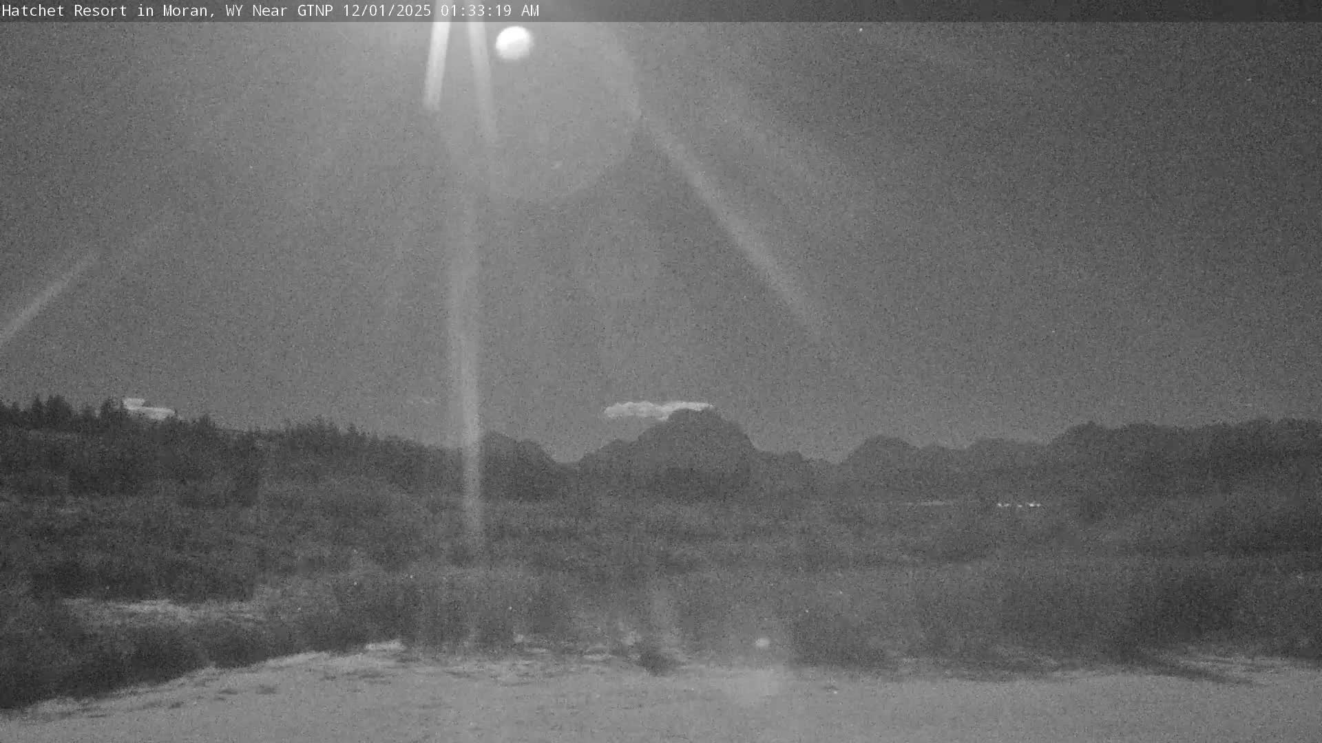 A grainy, monochromatic nighttime outdoor scene depicts a brilliant moon with radiating light rays in a clear sky over a vast landscape of dark vegetation in the foreground, silhouetted mountains topped by a small cloud, and distant faint structures.
