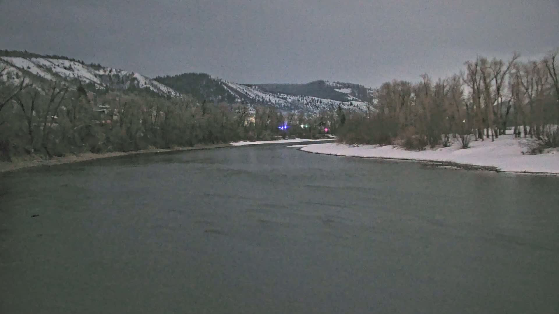 A wide, dark river flows past snow-covered banks lined with leafless trees under a cloudy, gray sky.