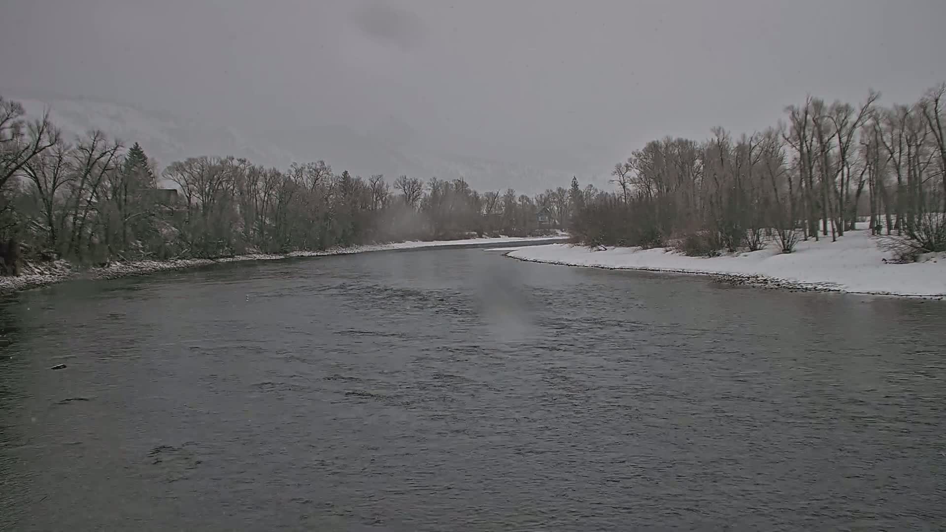 A wide, dark river flows between snow-covered banks lined with leafless trees under a gray, overcast sky.