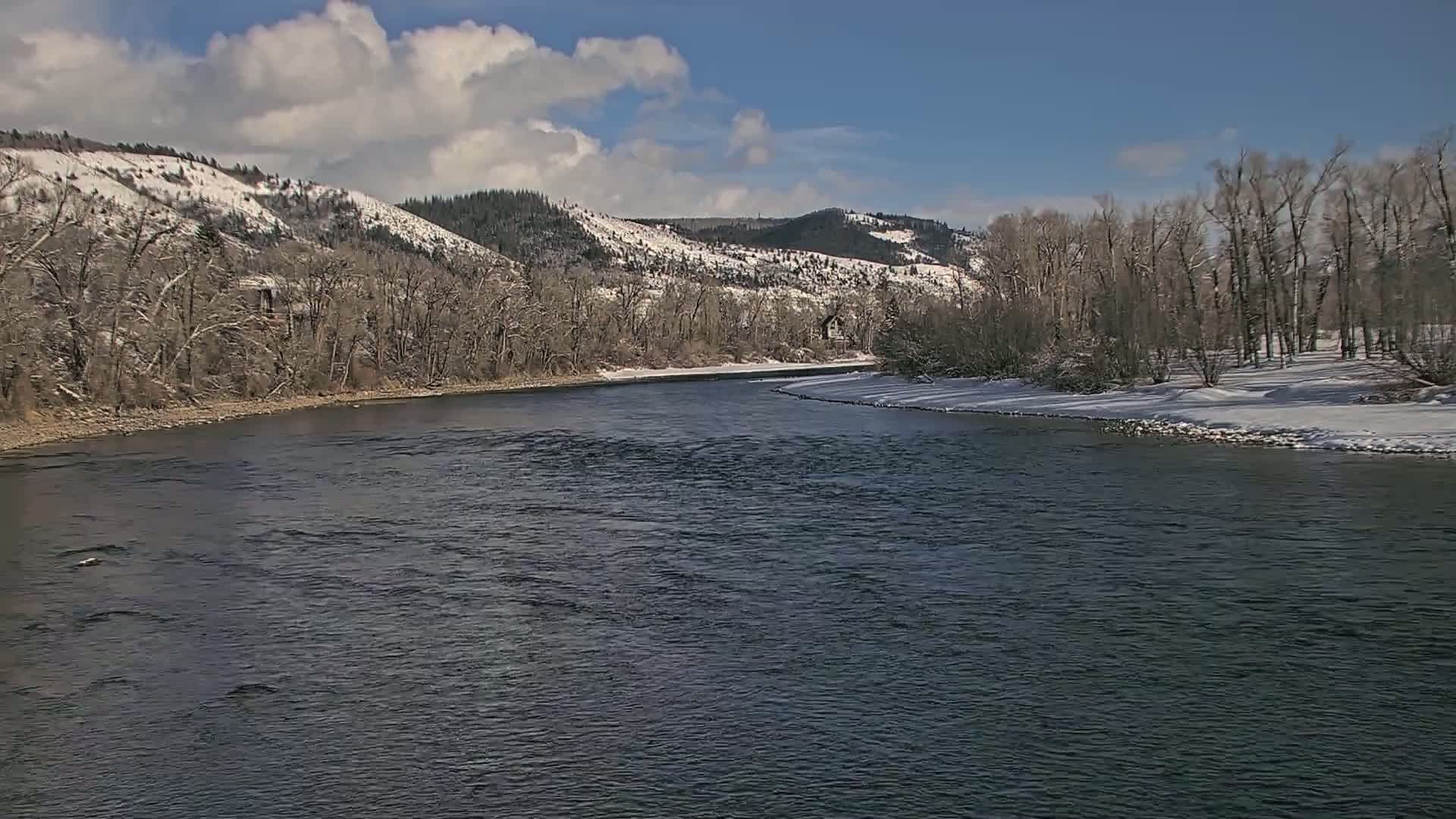 A dark river flows through a snow-dusted landscape under a partly cloudy sky.