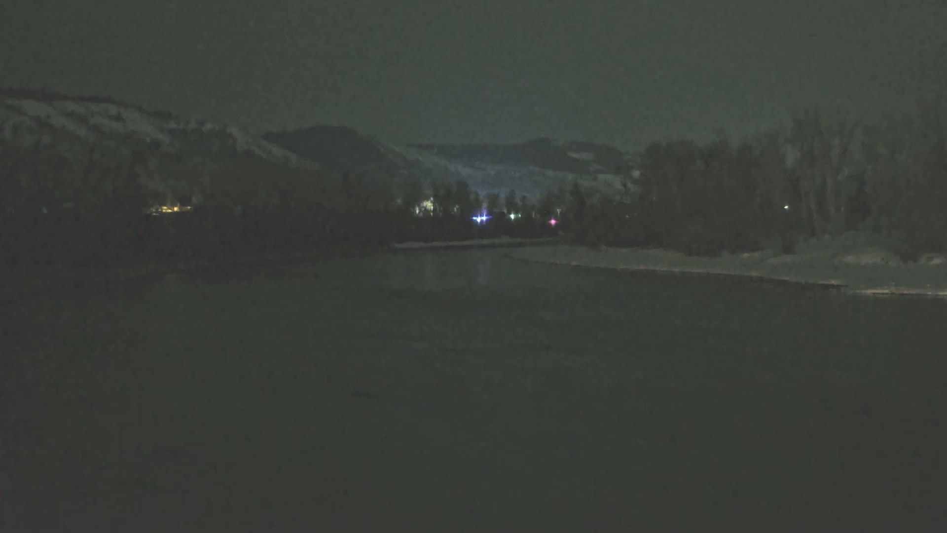A dark, nighttime scene shows a river in the foreground, with snow-covered banks and hills beyond, faintly lit by distant lights.