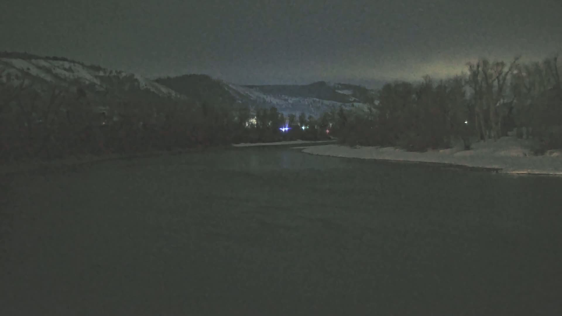 A dark, nighttime view of a river flowing between snow-covered banks, with dimly lit hills and trees in the background under a cloudy sky.