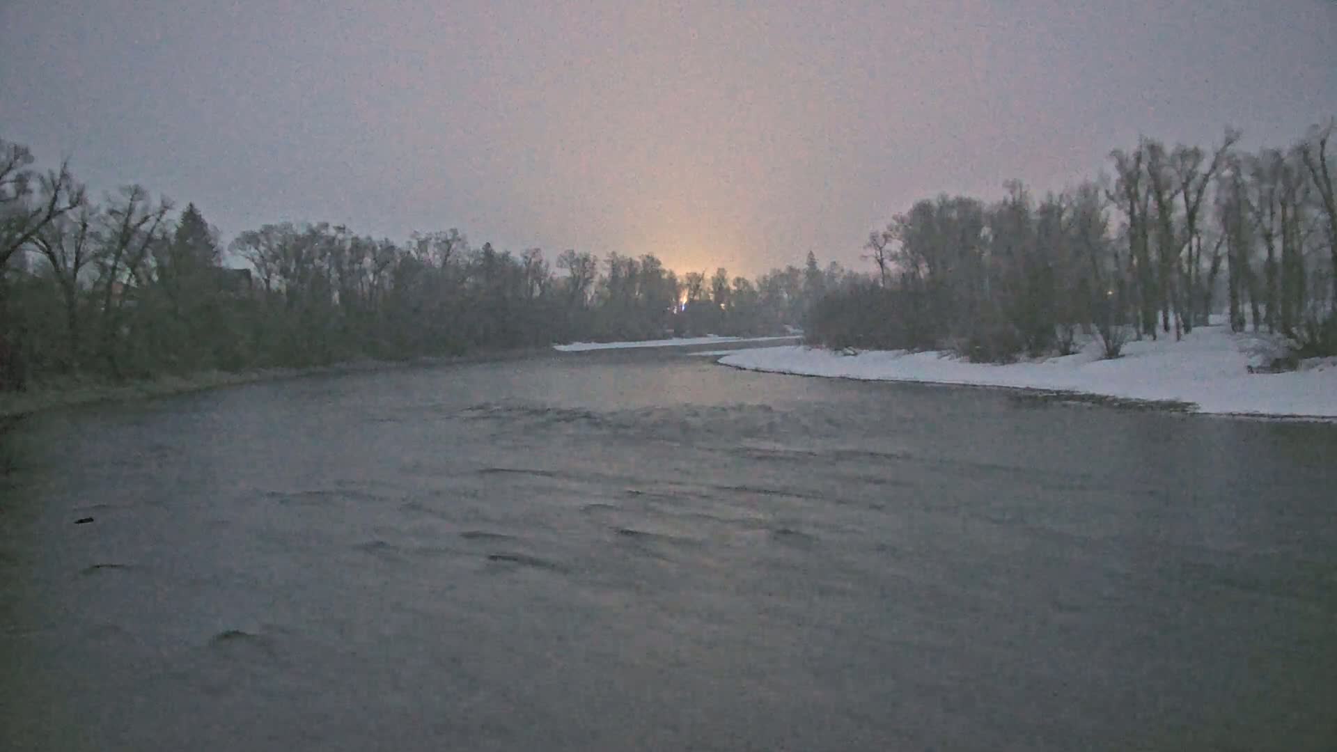 A gently flowing river curves past snow-covered banks lined with bare trees under a dusky, overcast sky.