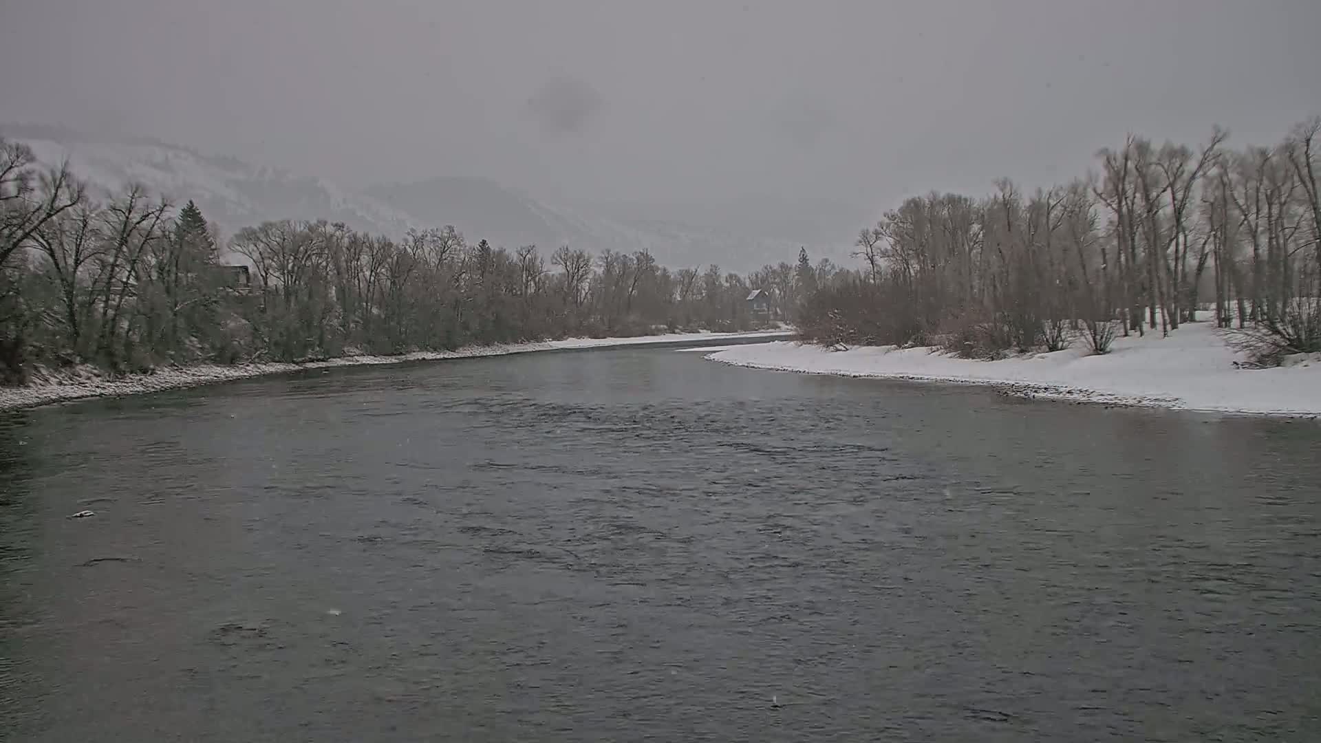 A snow-covered river bends gently through a landscape of snow-dusted trees and mountains under a gray, snowy sky.