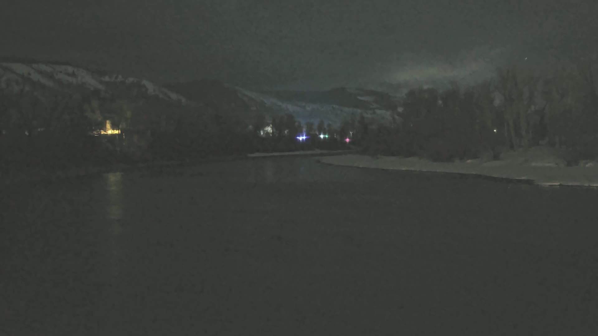 A dark, nighttime scene shows a calm body of water reflecting a few distant lights from a snow-covered hillside beyond a dark treeline.
