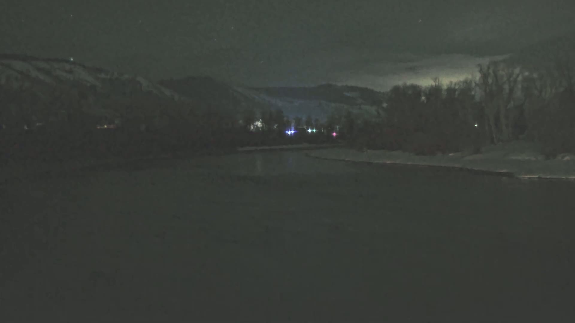 A dark, snow-covered landscape at night shows a river reflecting a few distant lights, with snow-covered hills and trees in the background under a partly cloudy sky.