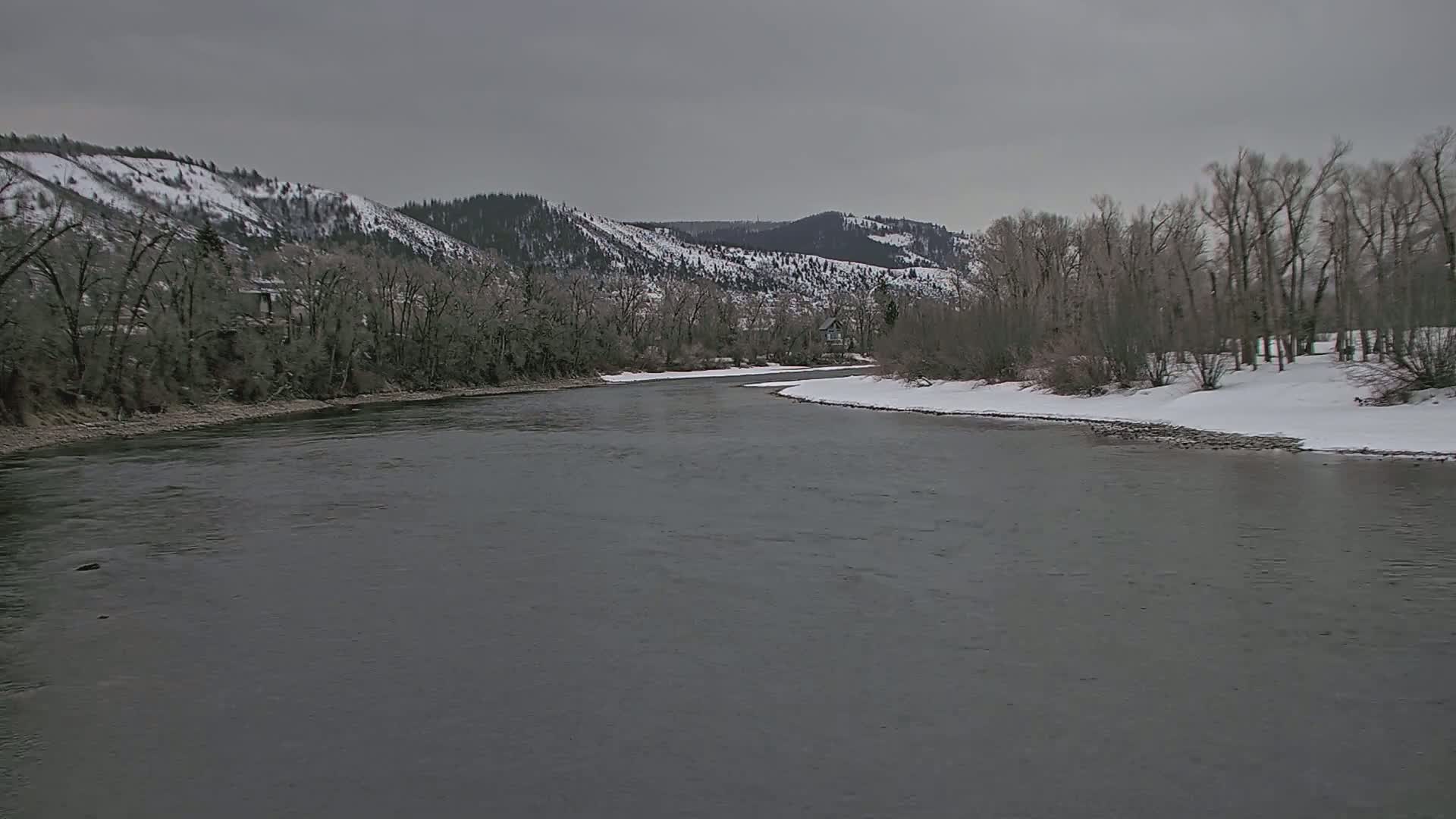 A wide, grey river flows past snow-covered banks lined with leafless trees and snow-dusted mountains under an overcast sky.