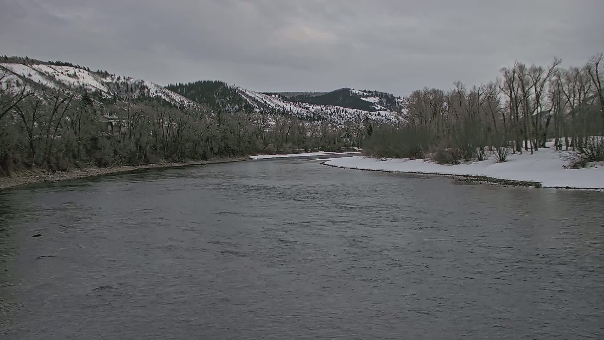 A wide, grey river flows past snow-covered banks lined with bare trees and snow-dusted mountains under an overcast sky.