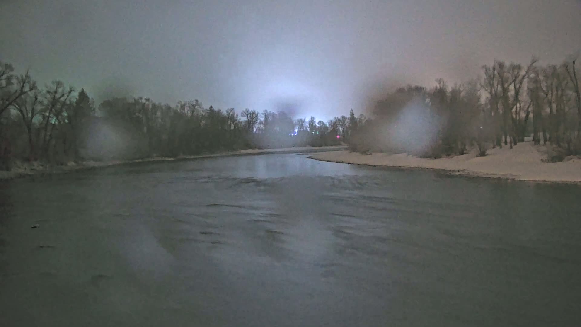 A dark, snow-covered riverbank with leafless trees is visible under a dusky, overcast sky.