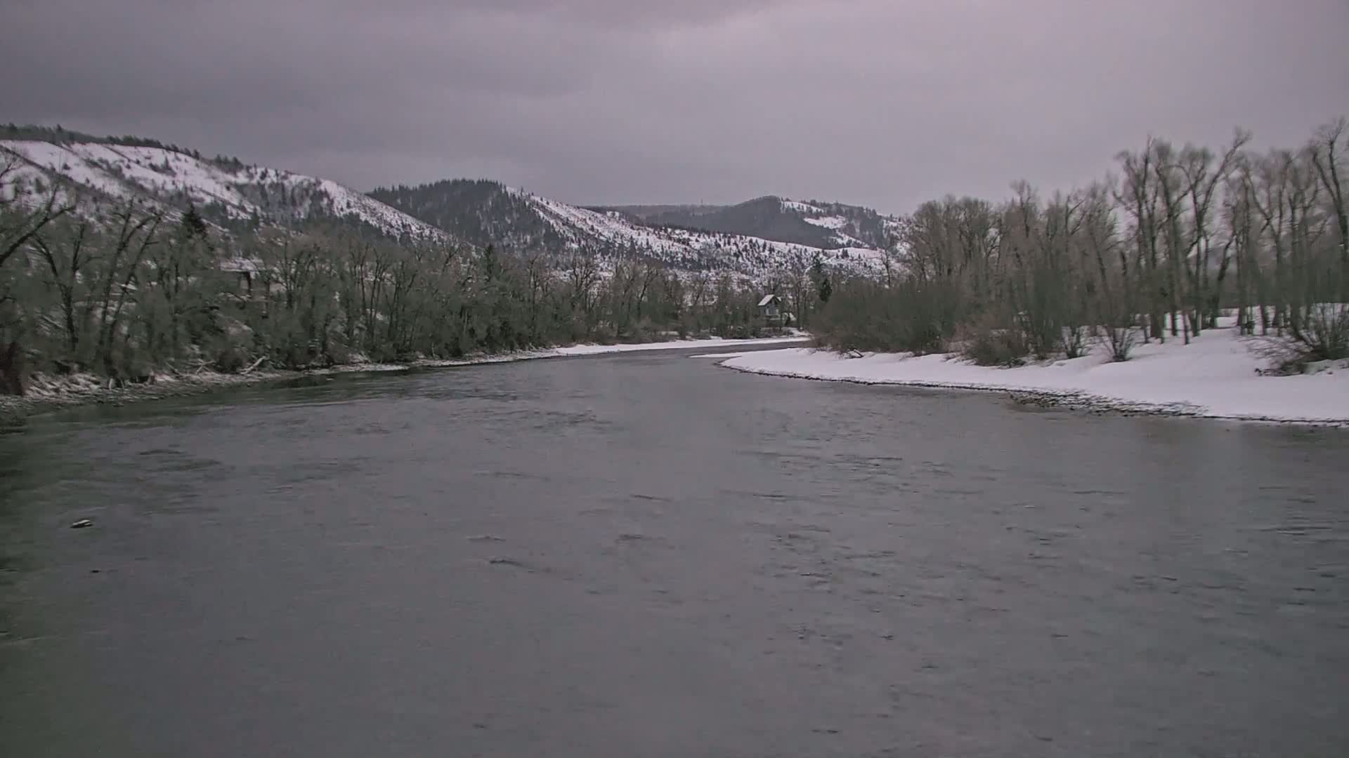 A wide, grey river flows past snow-covered banks lined with leafless trees and snow-dusted hills under an overcast sky.