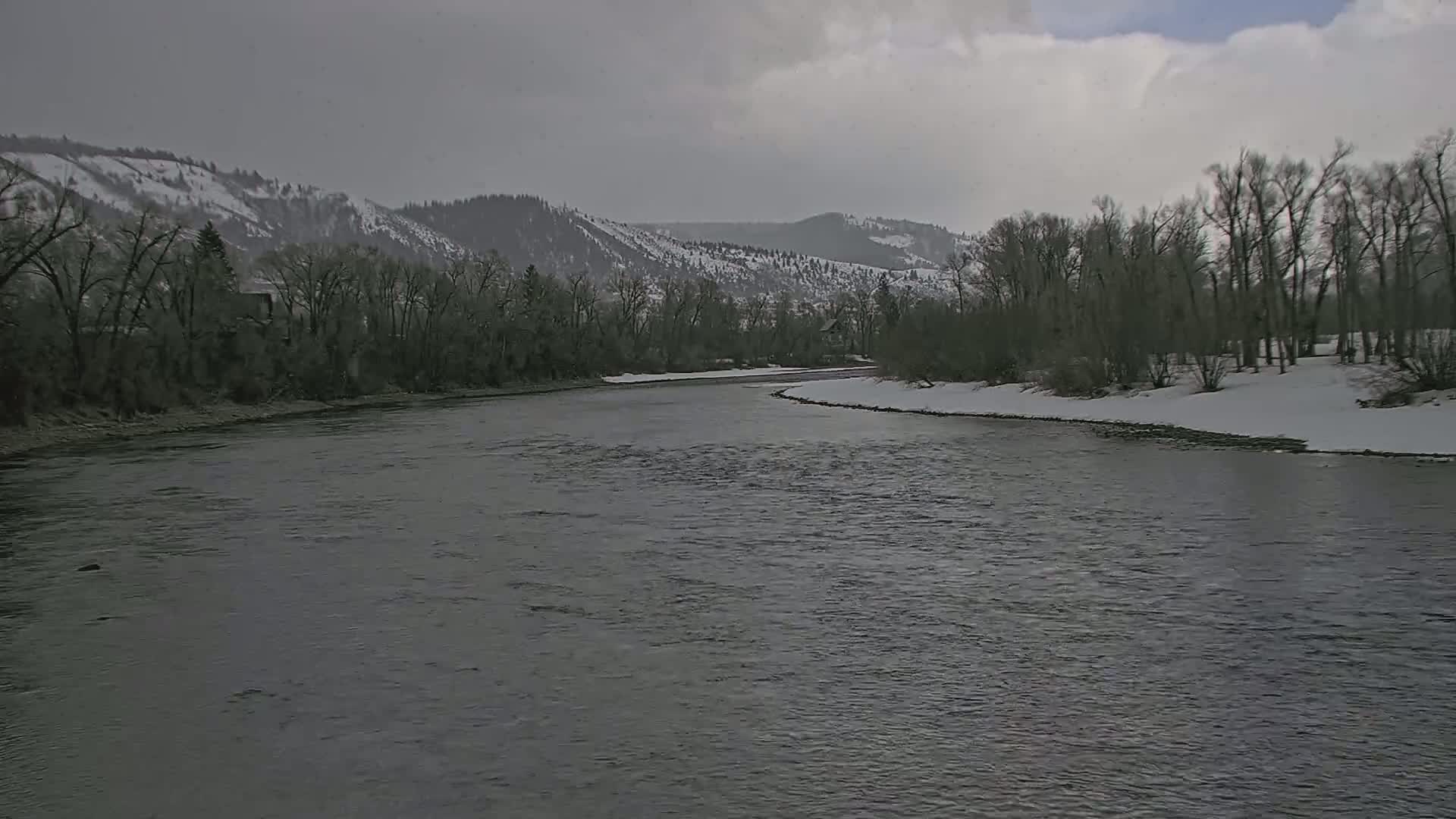 A wide, grey river flows through a snow-dusted landscape beneath a cloudy sky, with bare trees lining its banks and snow-covered mountains in the distance.