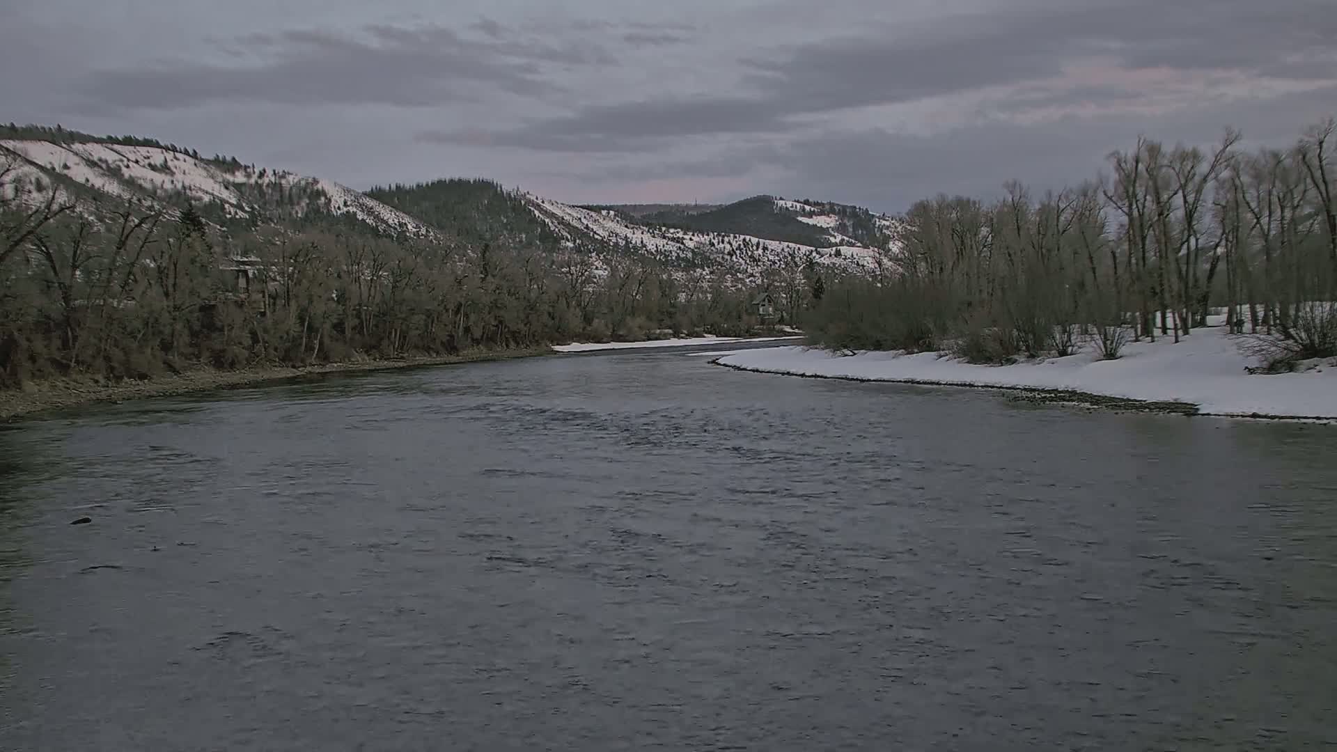 A wide, dark river flows past snow-dusted banks lined with bare trees under a cloudy sky, with snow-capped mountains in the background.