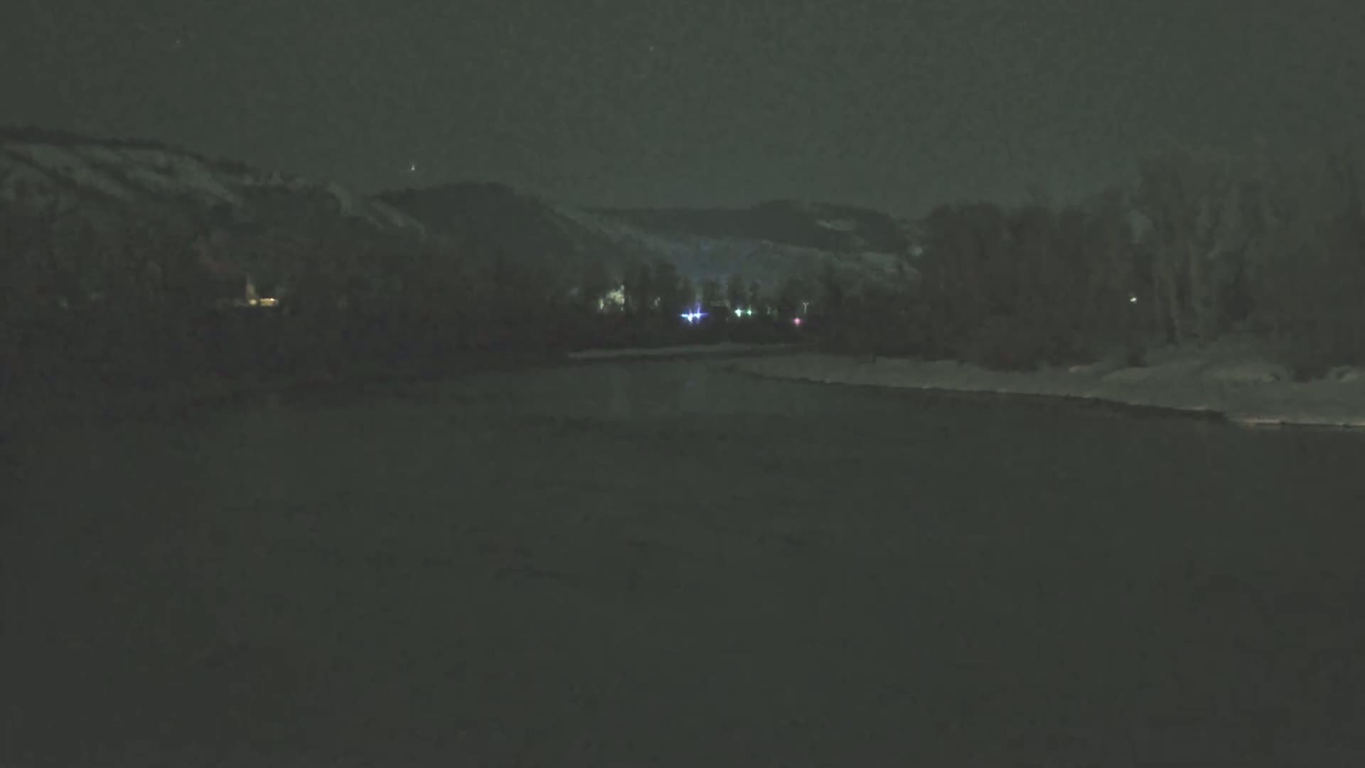 A dark, nighttime scene shows a calm body of water reflecting a few distant lights against a snow-covered bank and dark hills under a clear, starless sky.