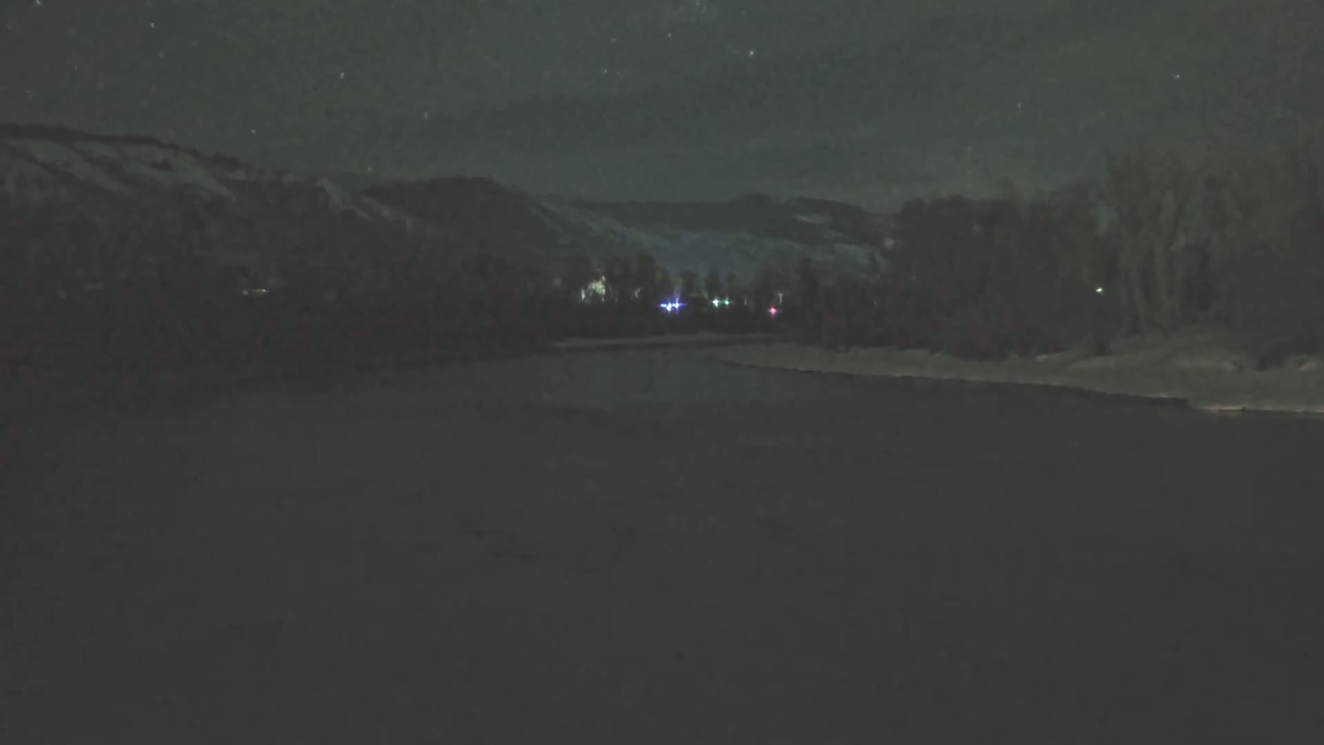 A snow-covered landscape at night is illuminated by distant lights and a starry sky.