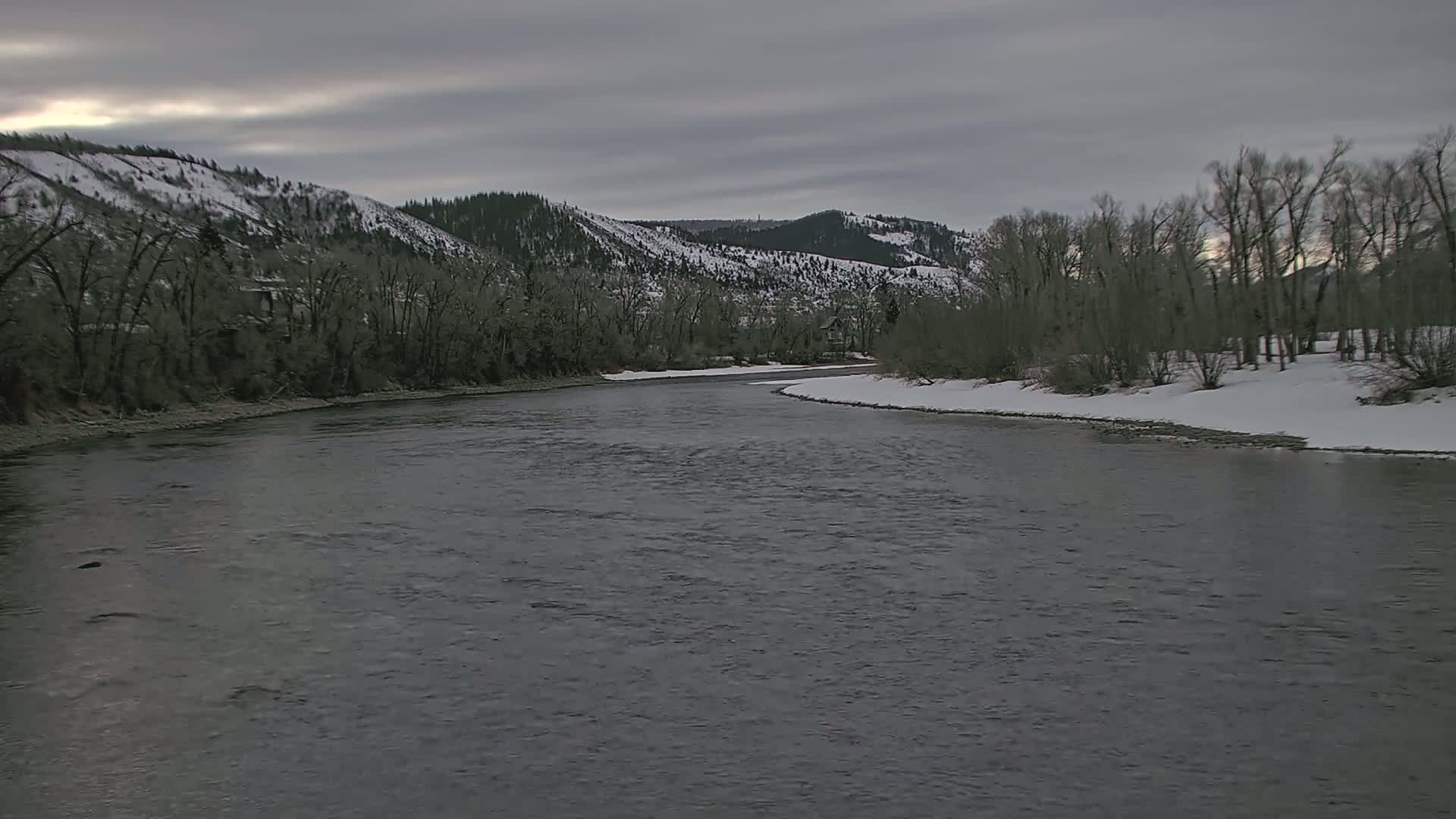 A dark-grey river meanders through a snow-dusted landscape under an overcast sky, past bare trees and snow-covered hills.