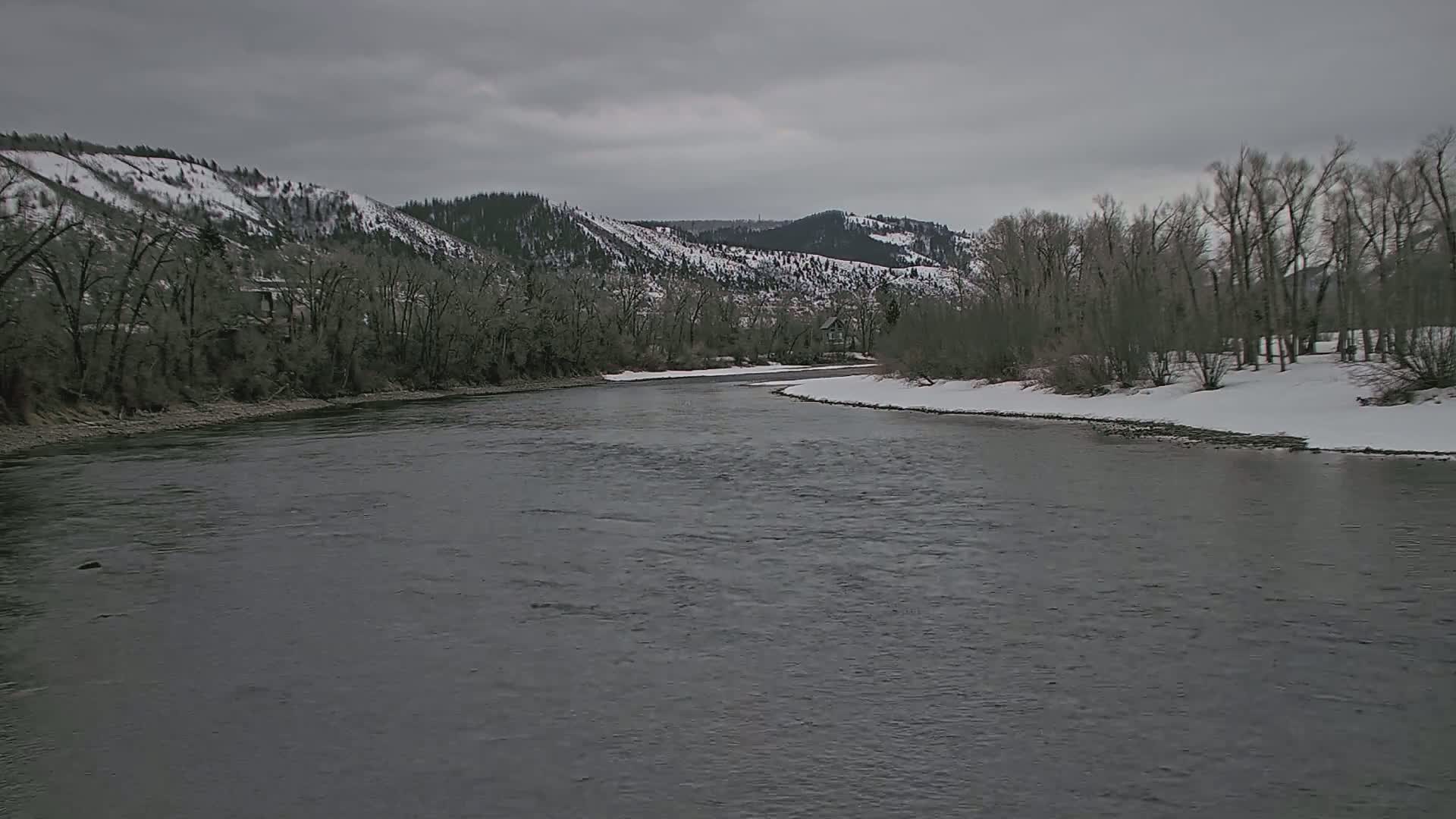 A wide, grey river flows past snow-dusted banks lined with leafless trees and snow-capped mountains under an overcast sky.