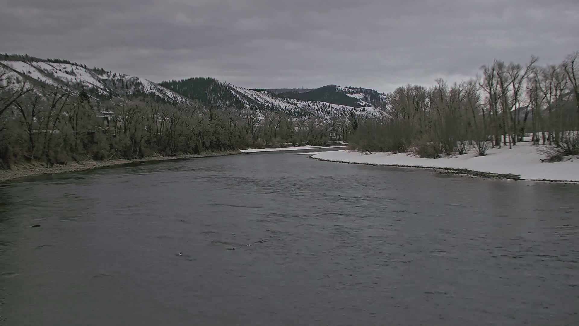 A wide, dark river flows past snow-covered banks lined with bare trees and snow-dusted hills under an overcast sky.