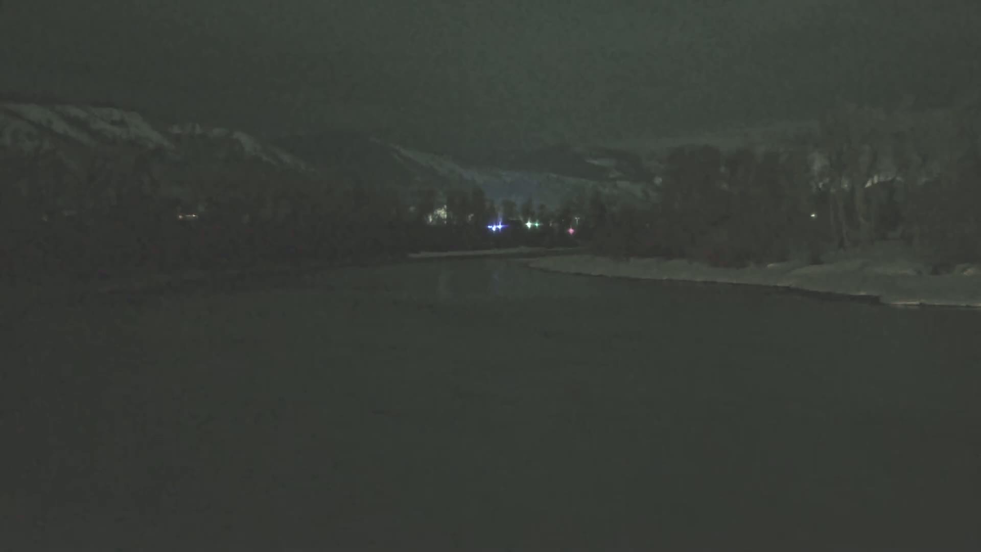 A dark, nighttime scene shows a snow-covered riverbank with faintly lit trees and distant mountains under a cloudy sky.