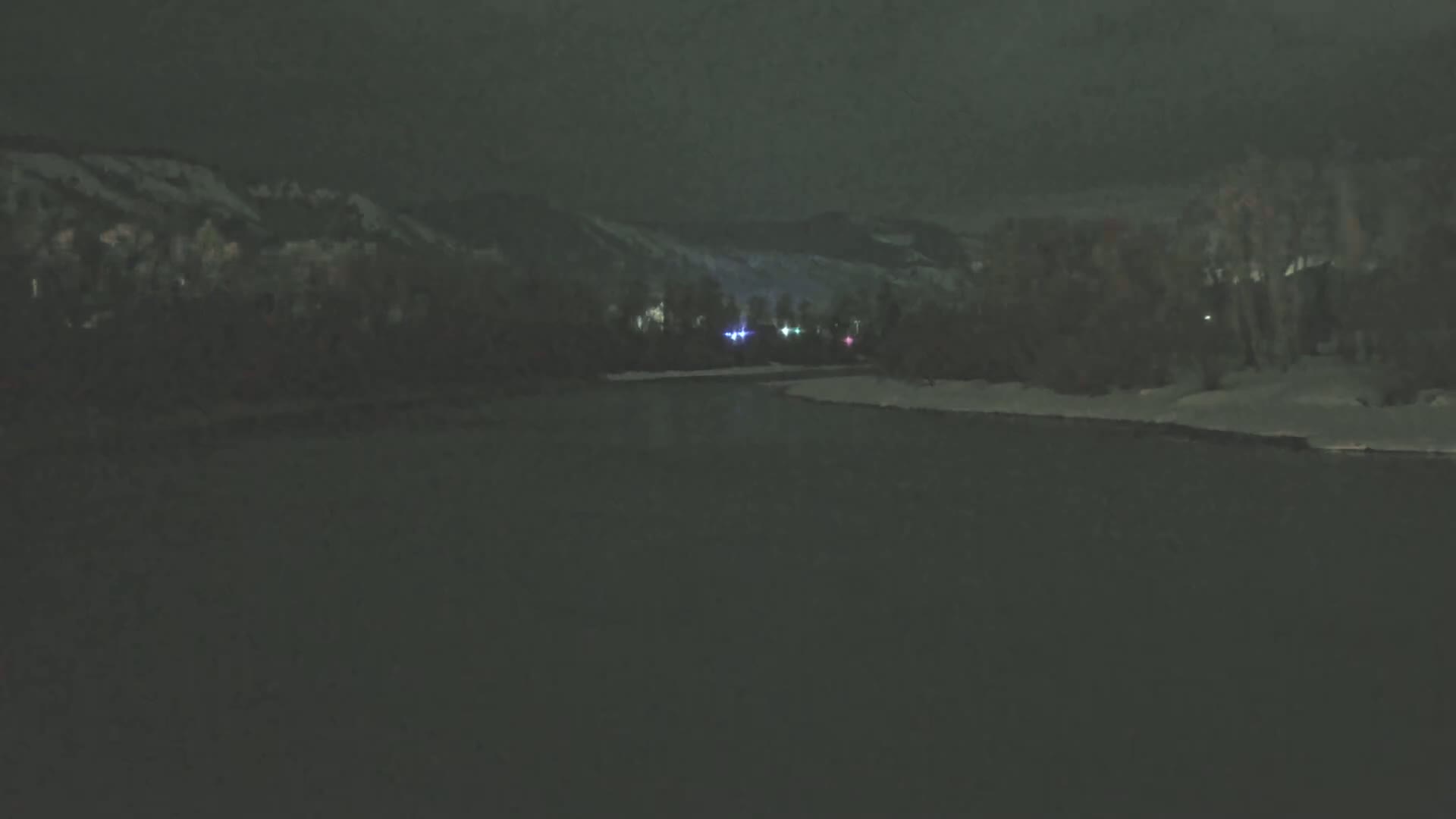 A dark, nighttime scene shows a snow-covered riverbank in front of snow-covered mountains under a cloudy sky.