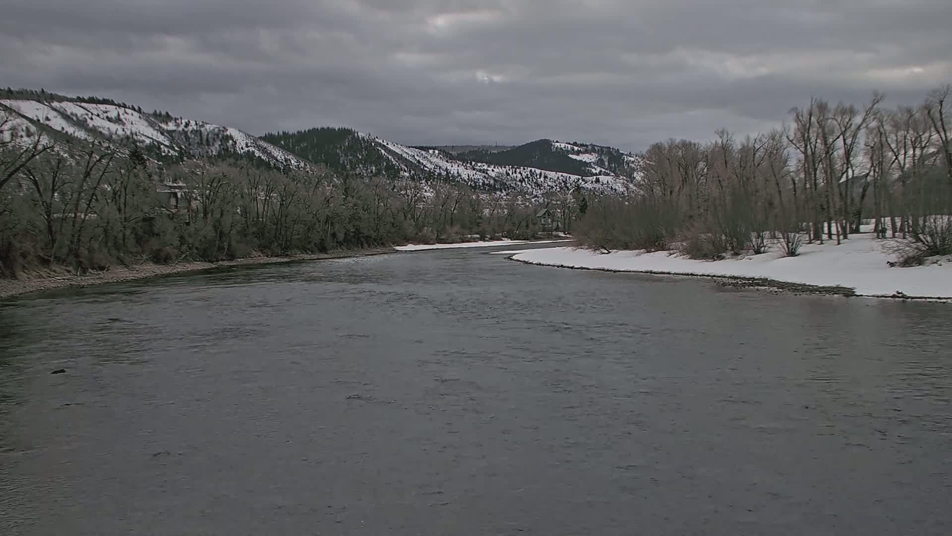 A wide, grey river meanders through a snow-dusted landscape under a cloudy sky, past bare trees and snow-capped hills.