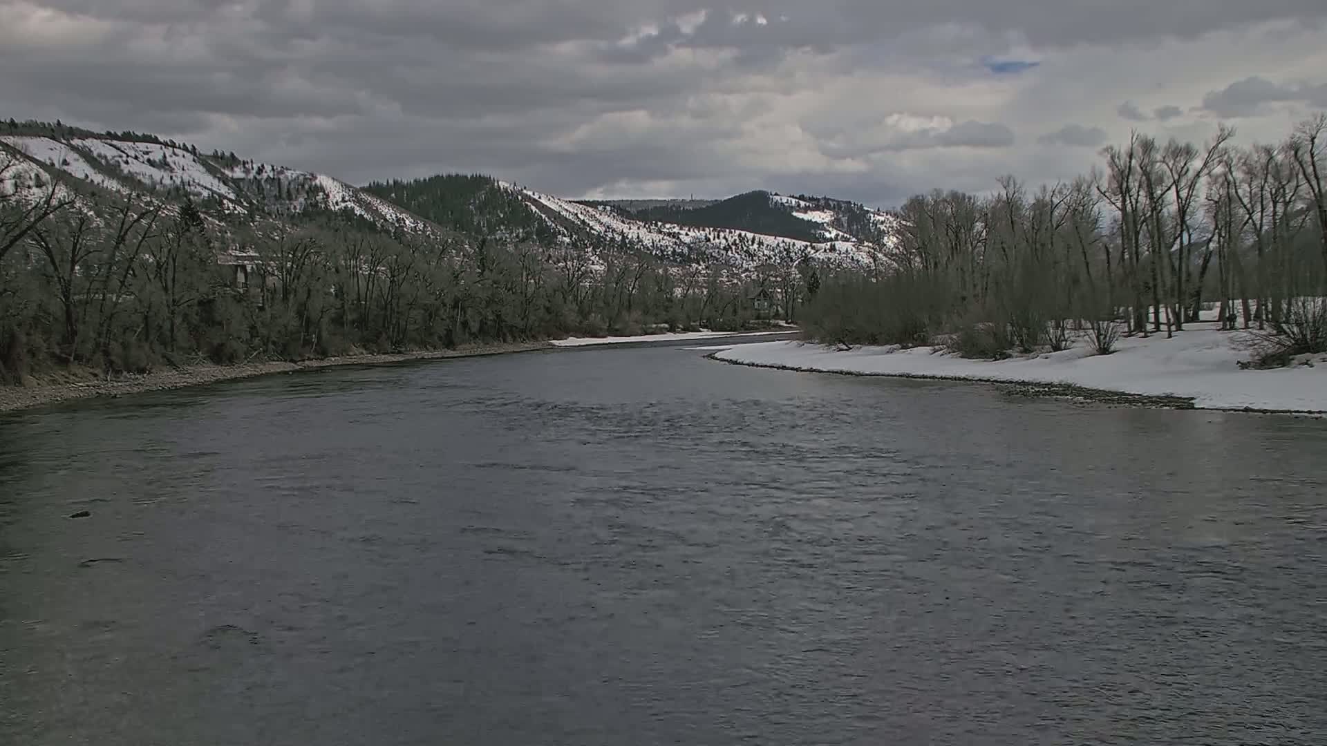 A dark river bends around a snow-dusted riverbank lined with bare trees under a cloudy sky, with snow-capped mountains in the background.