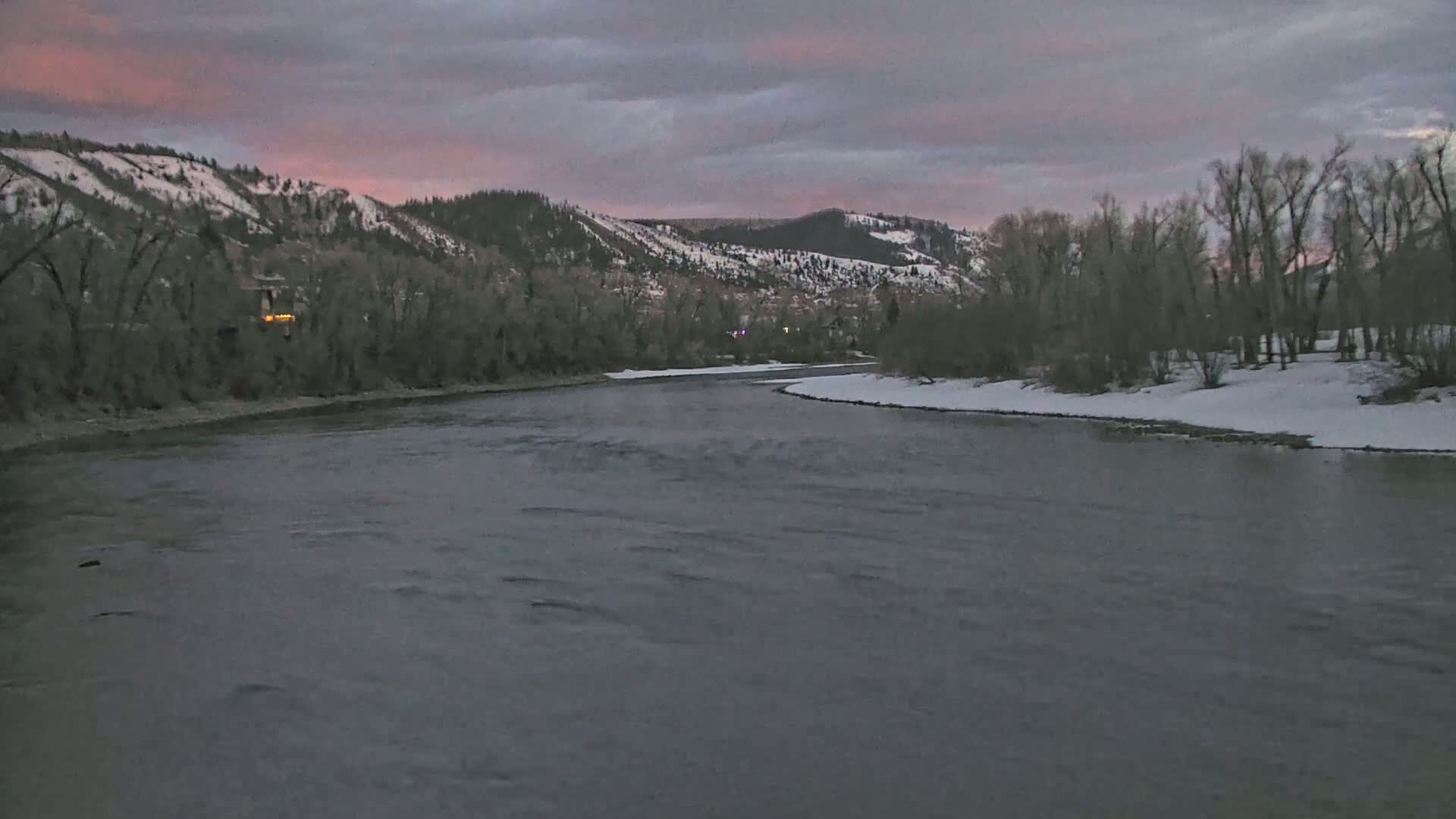A wide, gently flowing river under a dusky sky curves past snow-dusted riverbanks lined with bare trees and snow-capped hills in the background.