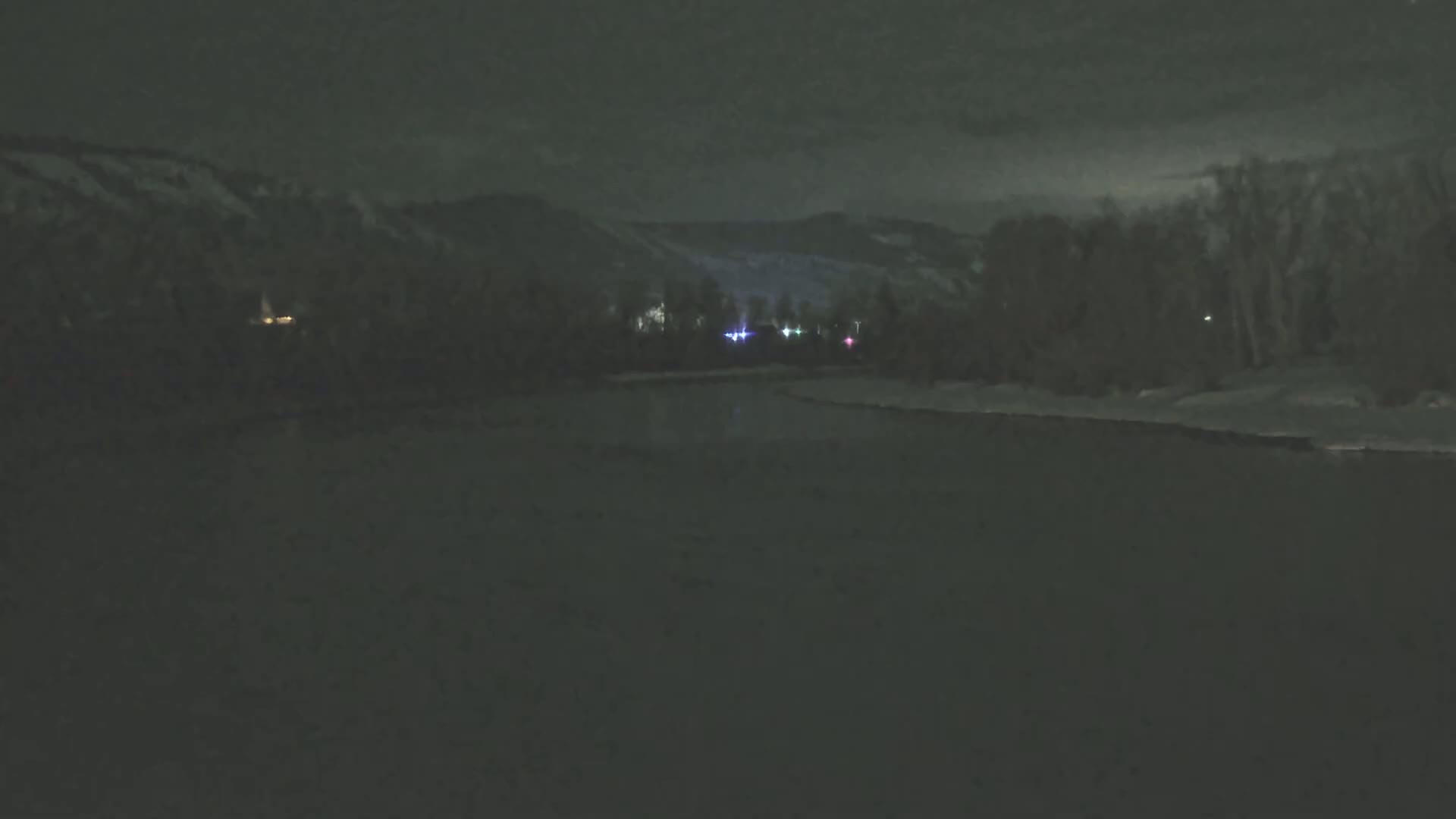 A dark, nighttime scene shows a body of water in the foreground, with faintly lit trees and snow-covered mountains in the background under a cloudy sky.