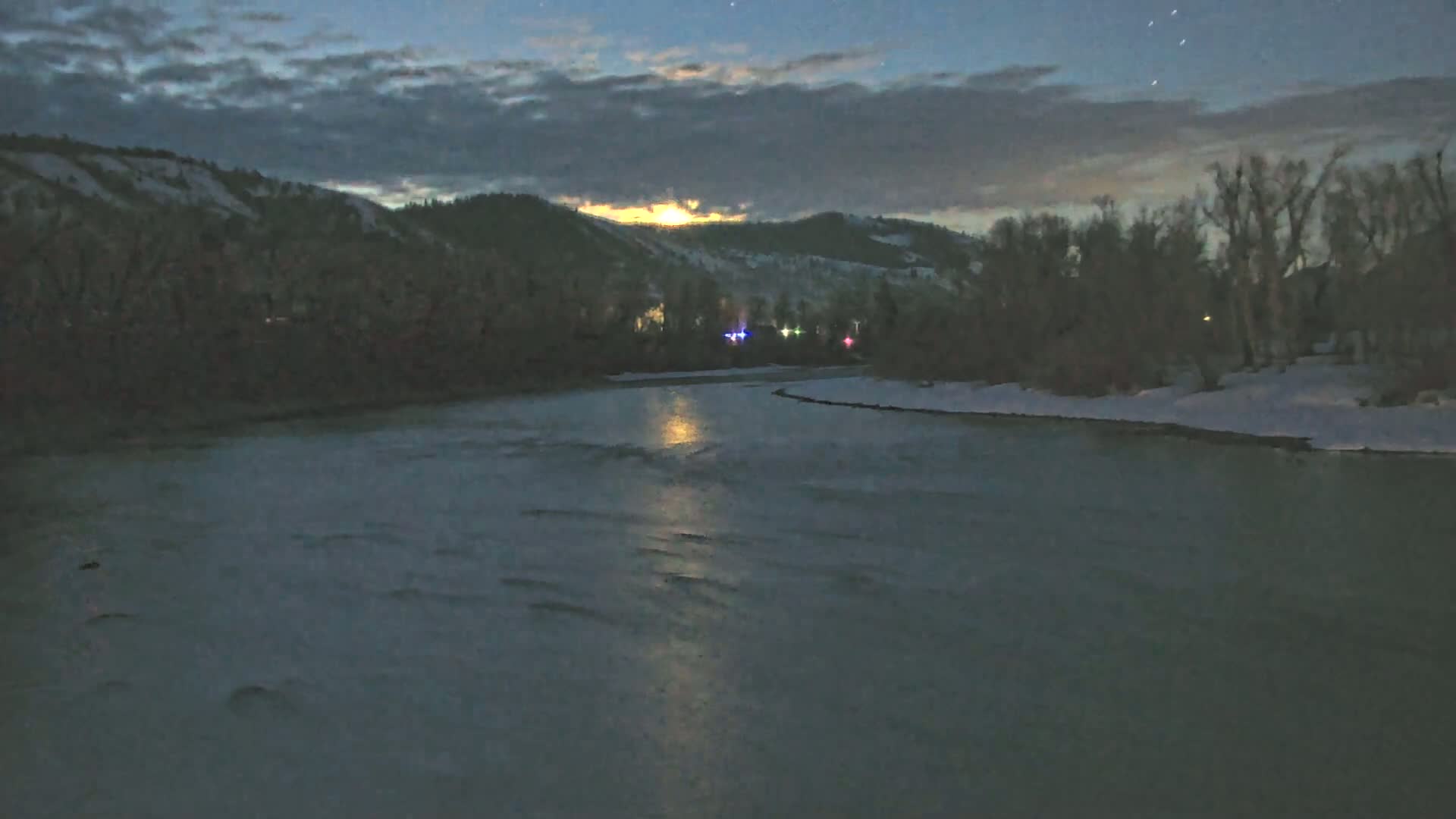 A moonlit river flows under a partly cloudy night sky past snow-covered banks lined with bare trees and hills in the distance.