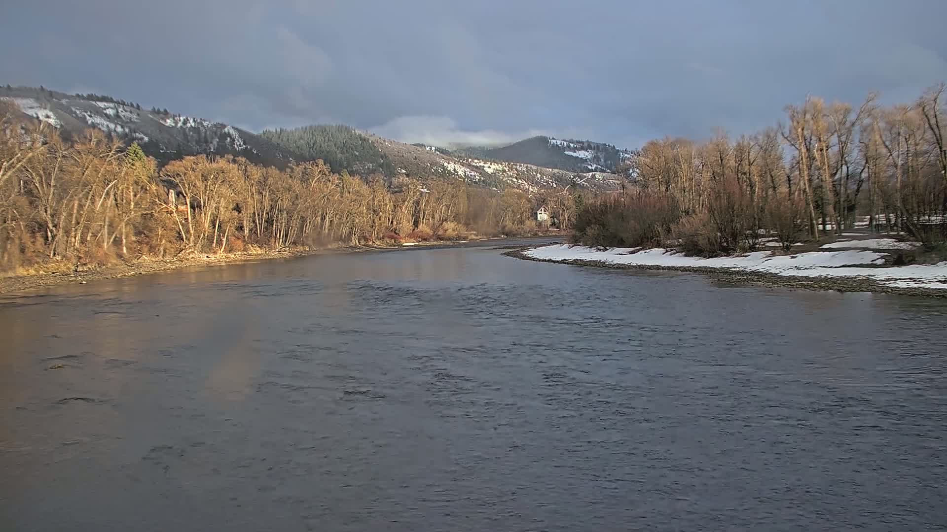 A wide, grey river flows past snow-dusted banks lined with bare trees under a partly cloudy sky, with snow-capped mountains in the background.