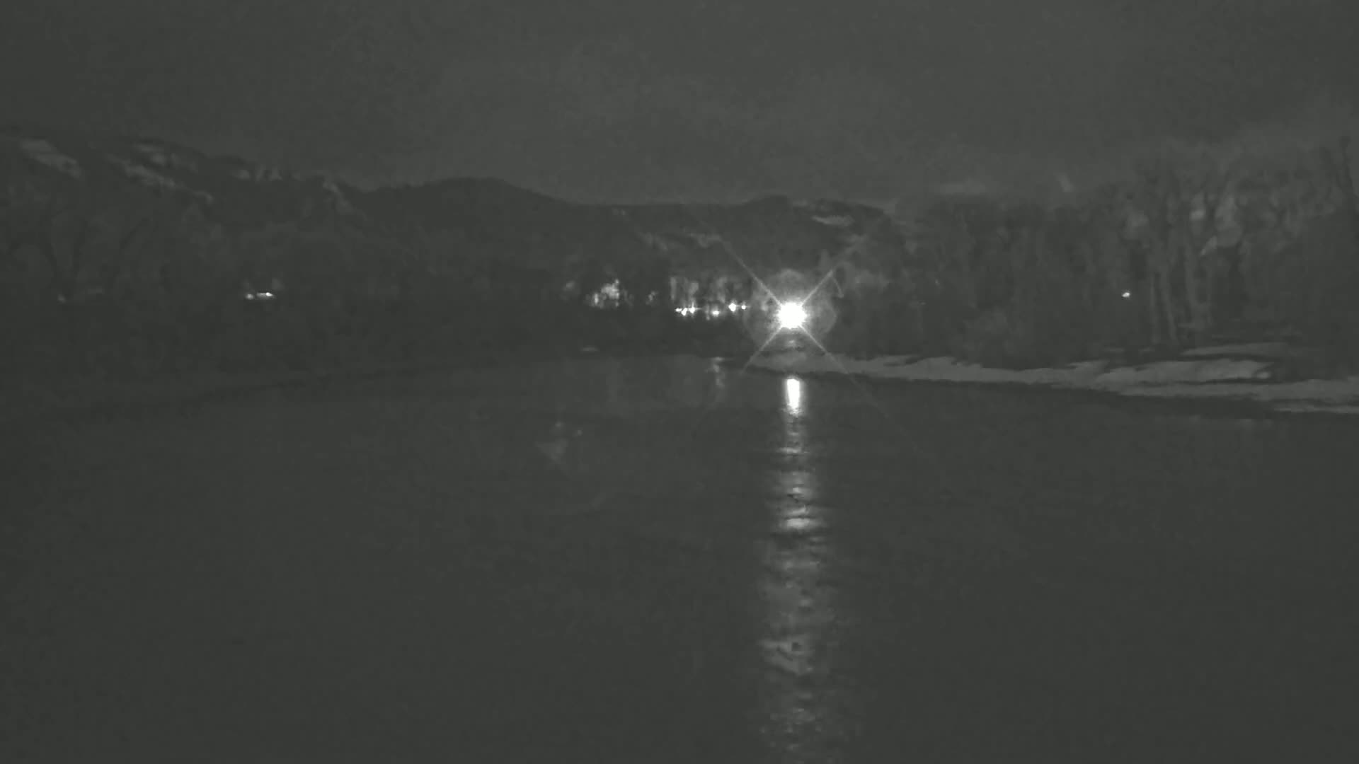 A dark, nighttime scene shows a calm body of water reflecting a distant light source, with a snow-covered shoreline and dark, mountainous silhouettes in the background.