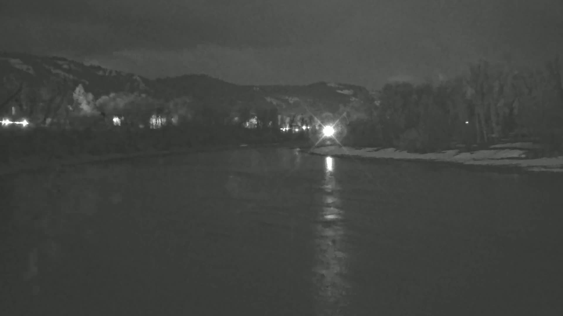 A dark, nighttime view of a river reflecting lights from the opposite bank, with snow-covered land and trees bordering the water under a mostly cloudy sky.