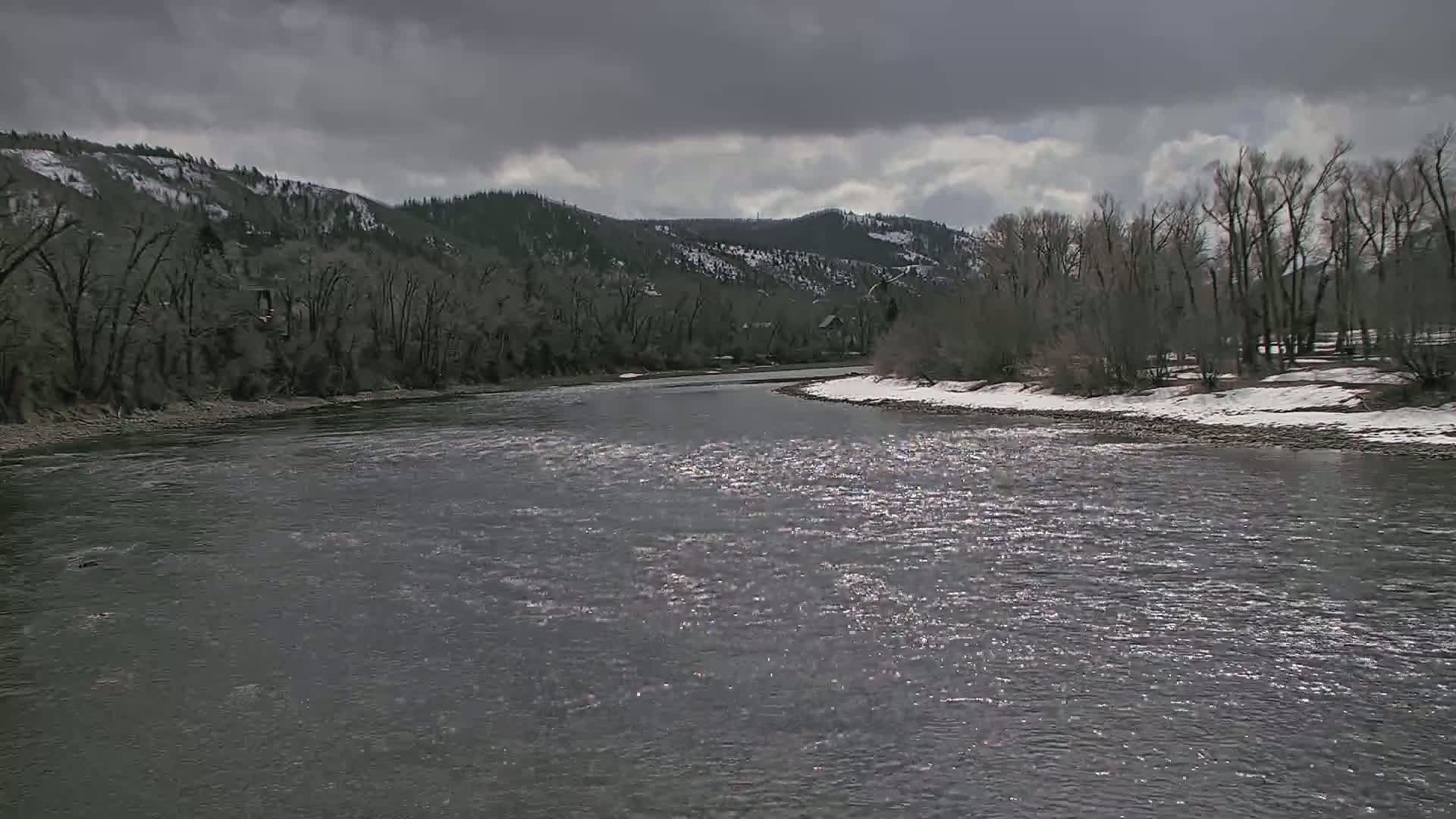 A wide, gray river flows under a cloudy sky past snow-dusted banks lined with bare trees and snow-capped hills in the distance.